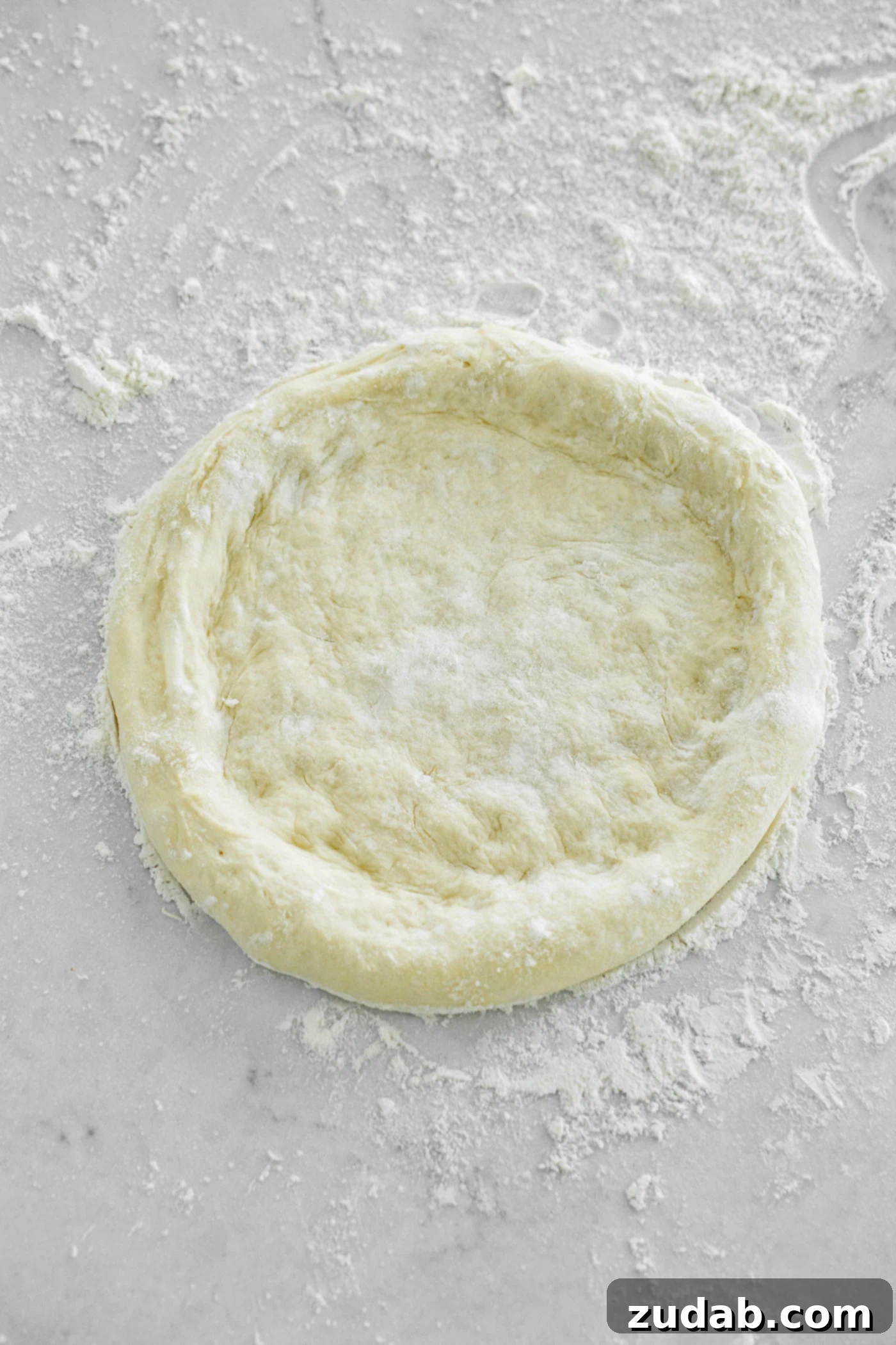 A freshly made, round homemade pizza dough on a floured surface, with beautifully raised edges ready for toppings, embodying a thick crust pizza style.