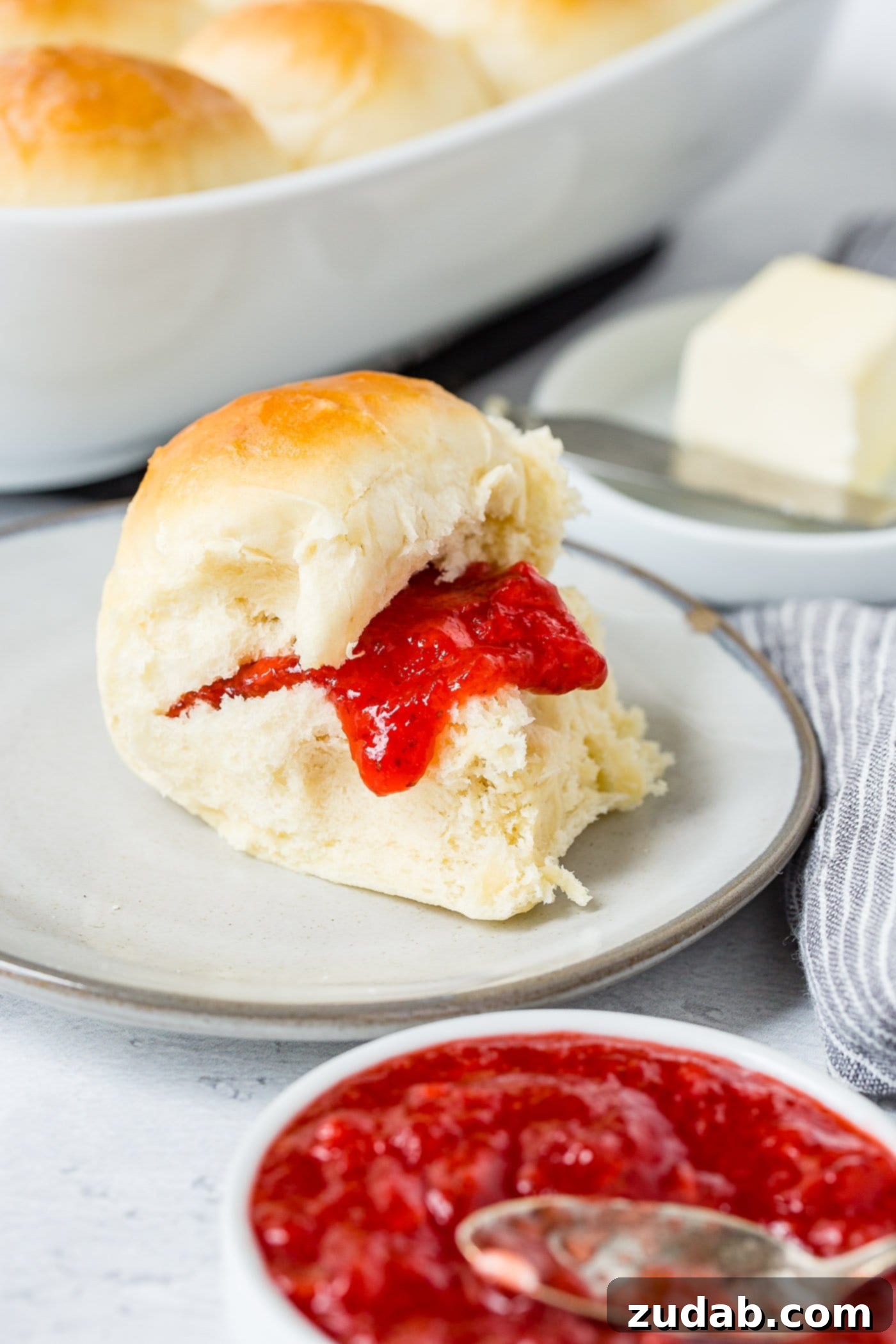 Dinner Rolls A white dinner roll on a small plate. There is strawberry jam on the roll. A dish of butter and a dish of jam are next to the roll.