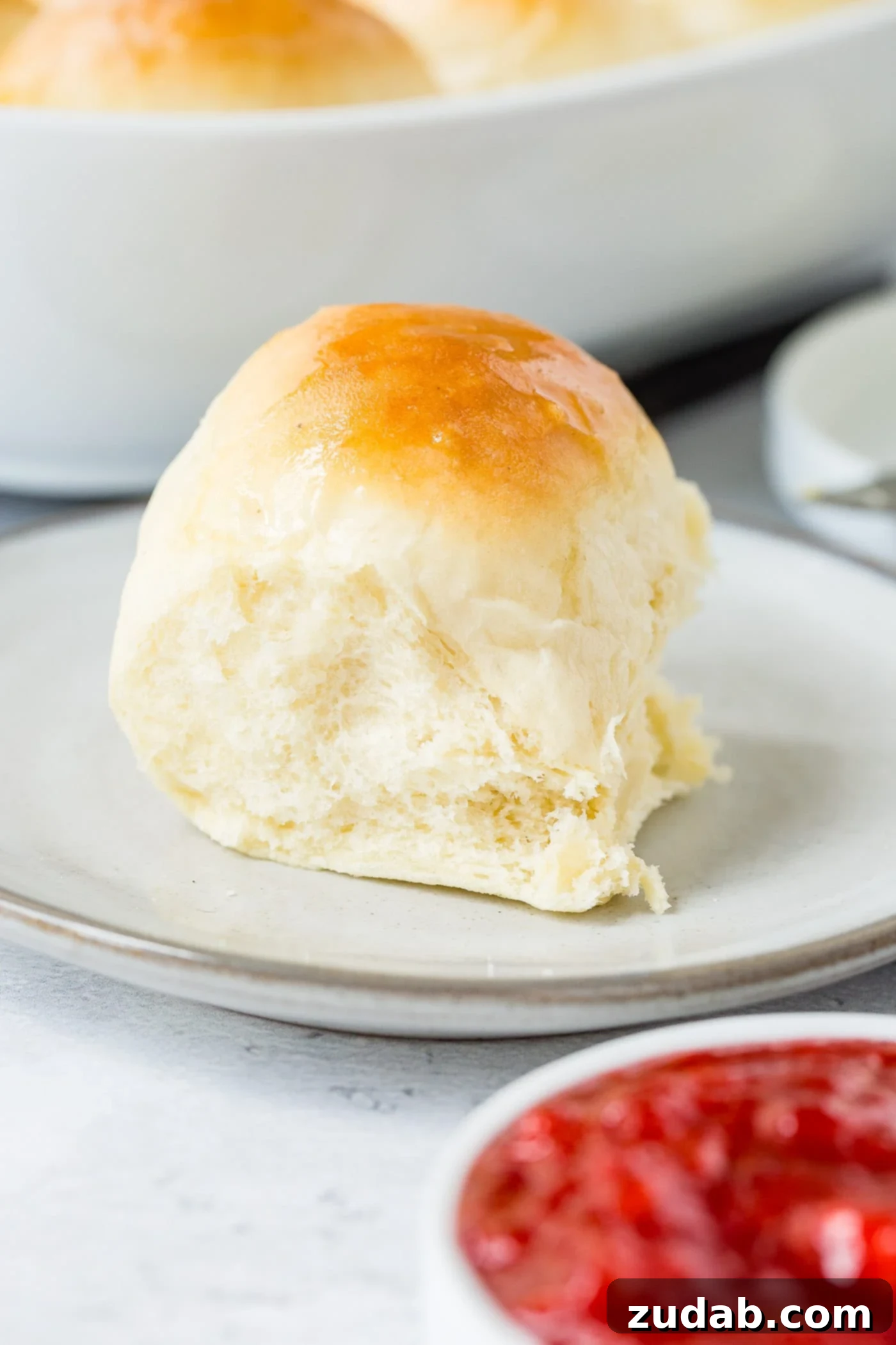 Dinner Rolls A white dinner roll on a small plate. The roll is perfectly baked to a golden brown. A small dish of strawberry jam is next to the roll.