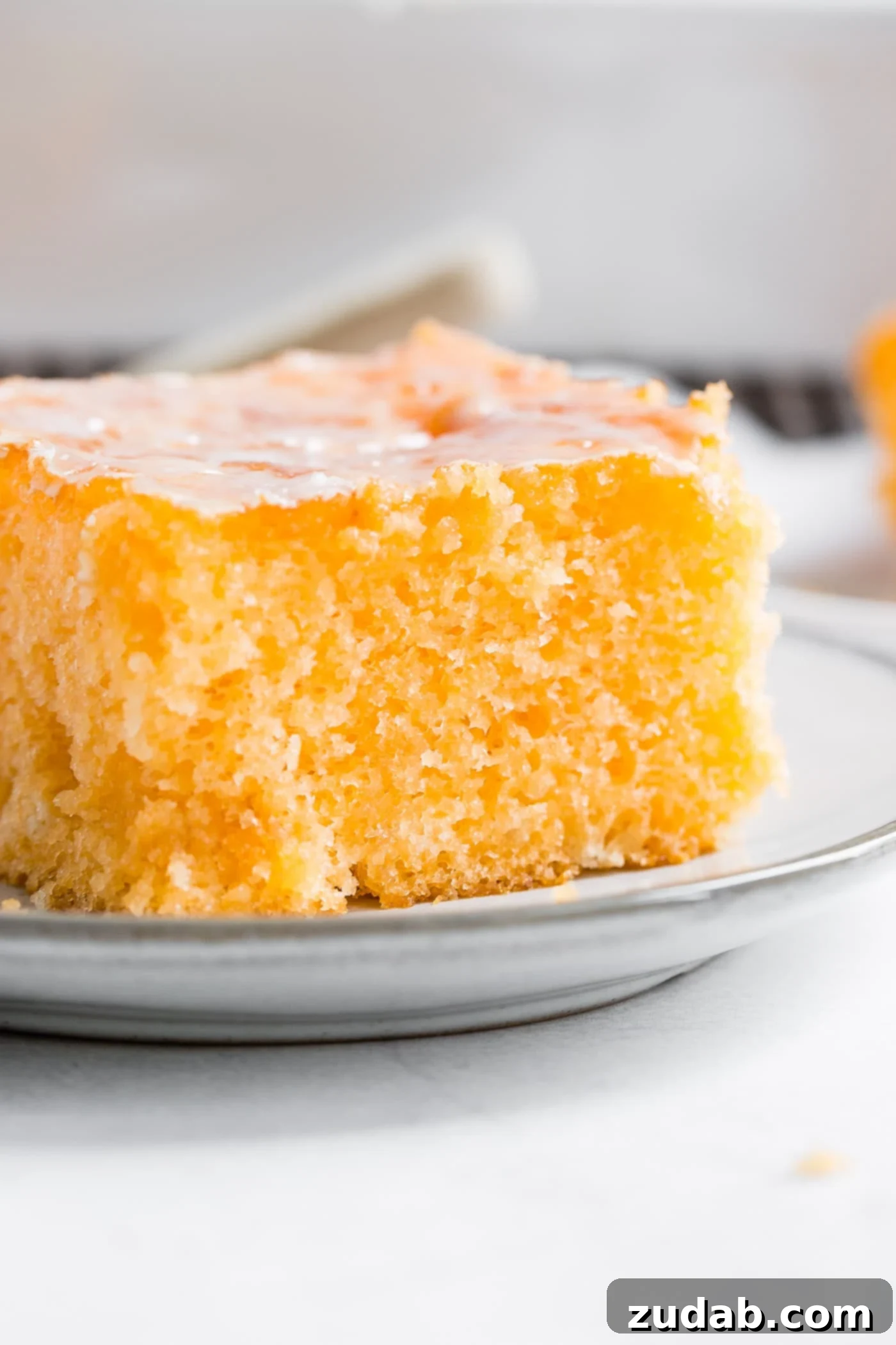 Zesty Orange Jello Cake Close-up A close-up photo of a piece of Orange Jello Cake on a small white plate, showing the soft texture and glaze.