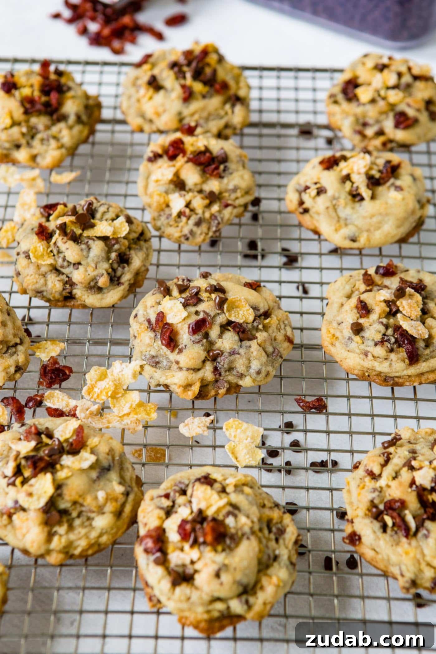 A close-up of Craisin Frosted Flake Chocolate Chip Cookies arranged on a wire cooling rack. Visible around and beneath the rack are scattered mini chocolate chips and dried craisins, emphasizing the delicious ingredients that make these cookies special.