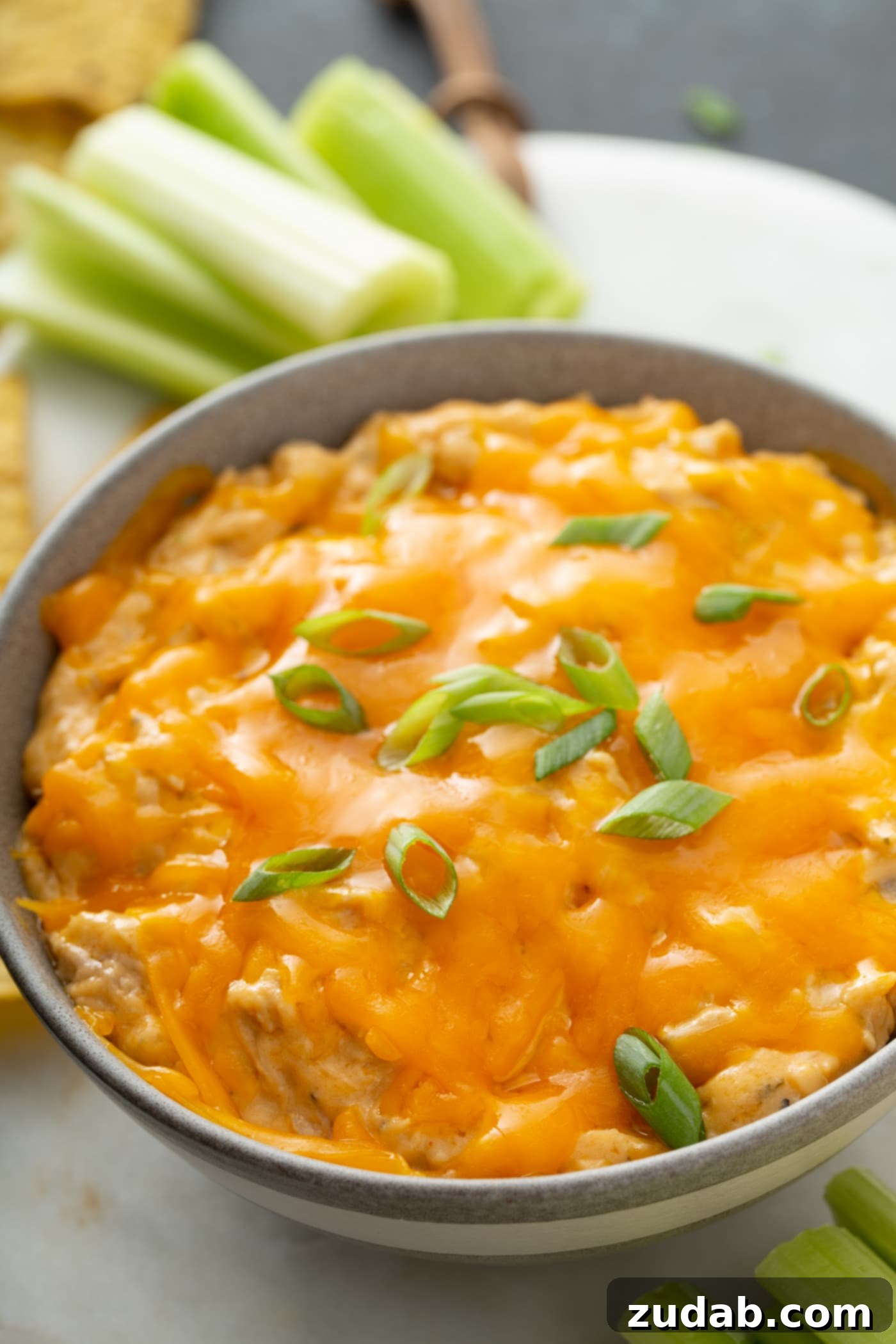 Slow Cooker Buffalo Chicken Dip Bowl of hot Slow Cooker Buffalo Chicken Dip topped with melted cheese and green onions, with celery sticks in the background.