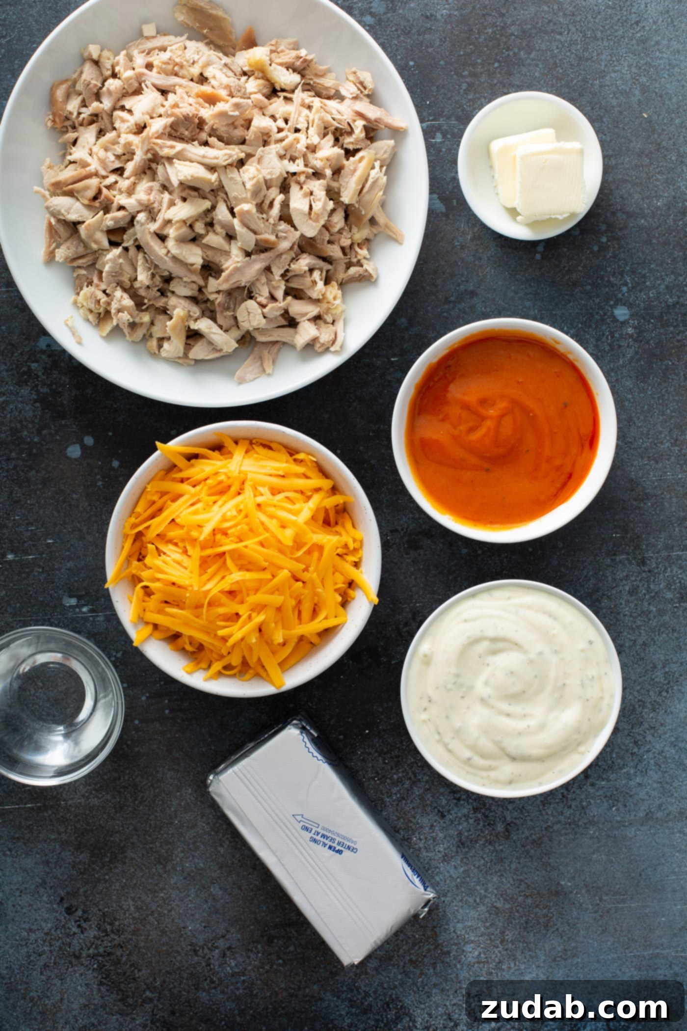 Slow Cooker Buffalo Chicken Dip Individual ingredients for Slow Cooker Buffalo Chicken Dip laid out: shredded chicken, cheddar cheese, cream cheese, buffalo sauce, ranch dressing, and water.