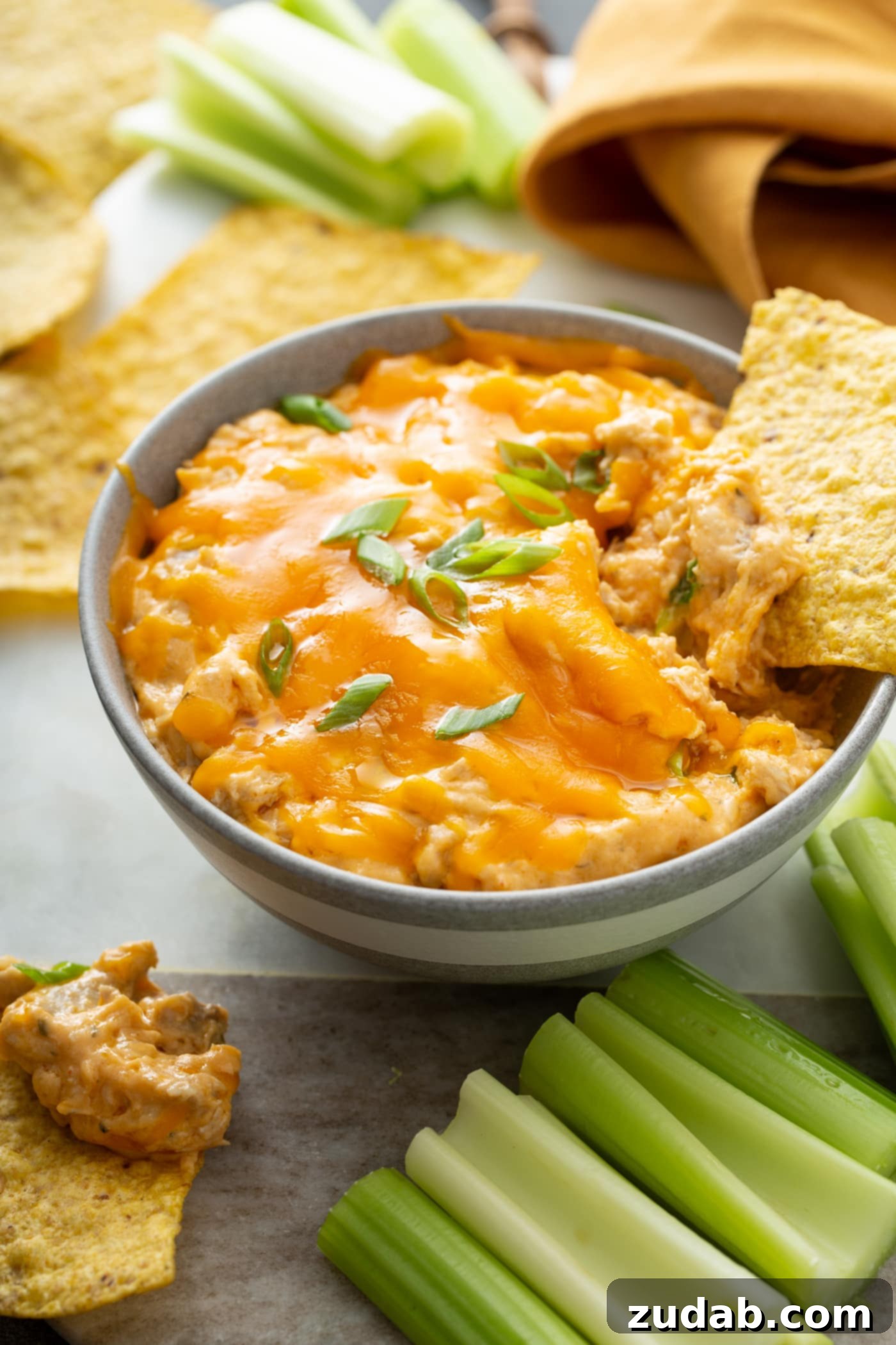 Slow Cooker Buffalo Chicken Dip Bowl of Slow Cooker Buffalo Chicken Dip garnished with green onions, surrounded by tortilla chips and celery sticks, with a chip being dipped.