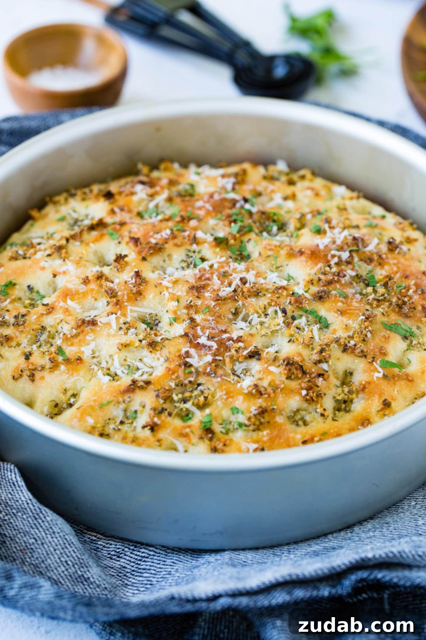 Garlic Parmesan Focaccia Bread A round baking dish with baked garlic parmesan focaccia bread. the bread is golden brown and has bits of green herbs in it. There are fresh herbs, measuring spoons and a small container of salt in the background.