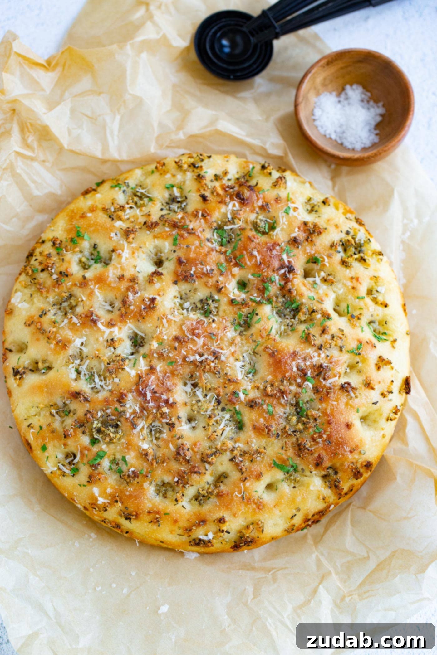 Garlic Parmesan Focaccia Bread Baked garlic parmesan focaccia bread sitting on parchment paper. It is baked to a golden brown with bits of green herbs on it. There is a small container of salt in the background.