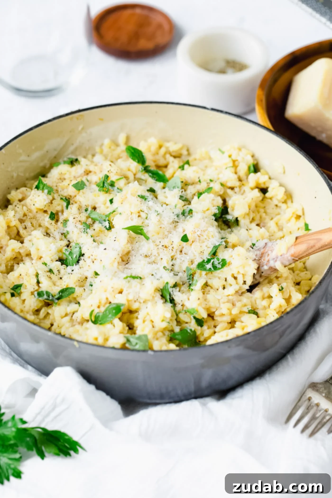 A bowl of creamy garlic parmesan orzo. It has a creamy white sauce and freah thyme leaves over it. There is a fork next to it and small containers of ingredients in the background. The bowl is sitting on a white tea towel.