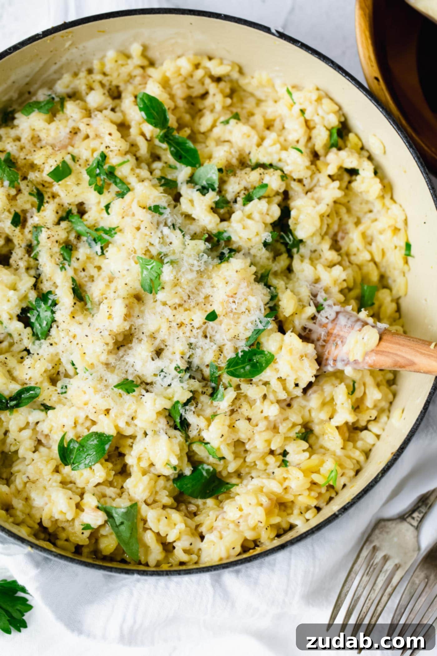 Creamy garlic parmesan orzo in a serving dish. There is a wooden spoon in the pasta. The pasta has been mixed with a creamy white sauce and fresh thyme leaves. Two forks are laying beside the bowl of pasta.