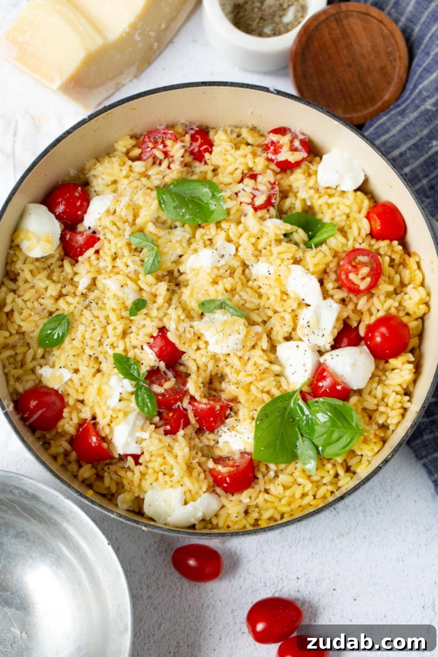 Caprese Orzo Pasta A vibrant bowl of Caprese Orzo Pasta salad, showcasing a delightful mix of ingredients. Prominently featured are bright red cherry tomatoes, creamy white mozzarella chunks or pearls, and verdant fresh green basil leaves, all perfectly mingled with the small, rice-shaped orzo pasta. The salad appears fresh and inviting, ready to be enjoyed as a light and flavorful meal.