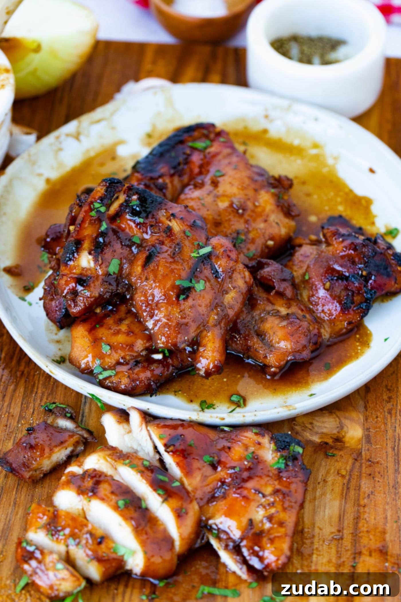Marinated Smoked Chicken Thighs on the Traeger A serving plate filled with juicy marinated smoked chicken thighs. Chopped parsley is sprinkled on top of the chicken and a sliced chicken thigh is on the cutting board next to the plate of chicken.