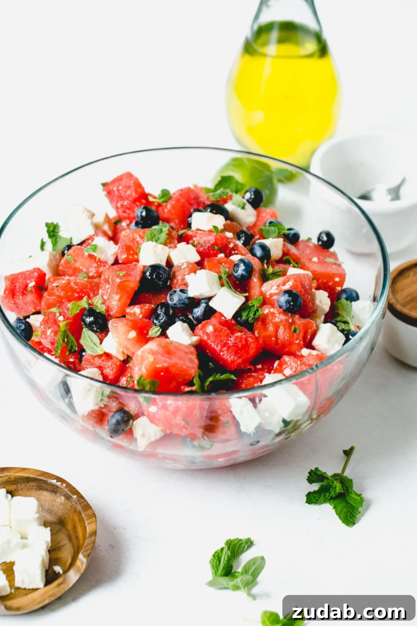 Blueberry Feta Watermelon Salad A glass bowl of blueberry feta watermelon salad. There are chunks of red watermelon, blueberries, and cubes of feta cheese. A few basil and mint leaves are sprinkled in the salad.