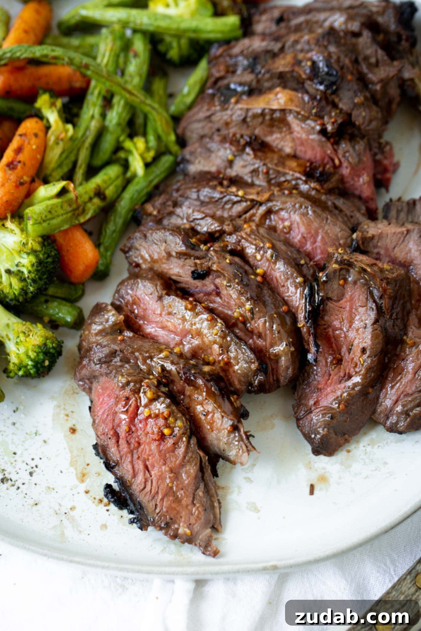 A photo of sliced medium rare hanger steak on a white plate next to a pile of roasted broccoli, baby carrots and green beans.