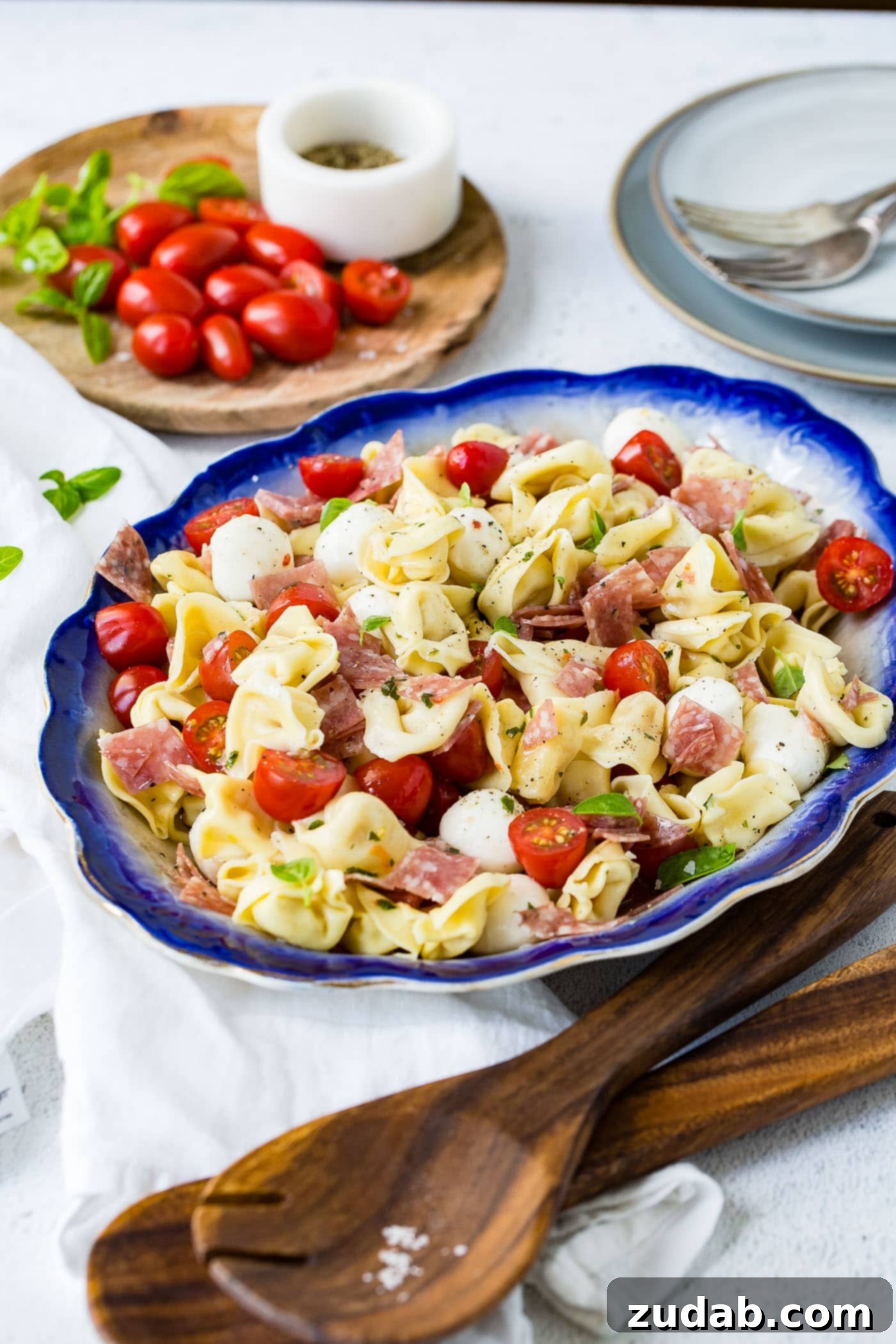 A blue-edged serving dish of 5-minute Italian salami tortellini salad. There are chunks of savory salami, vibrant slices of grape tomatoes, and creamy mozzarella balls nestled amongst the tender tortellini. A rustic cutting board adorned with fresh grape tomatoes provides a charming backdrop.