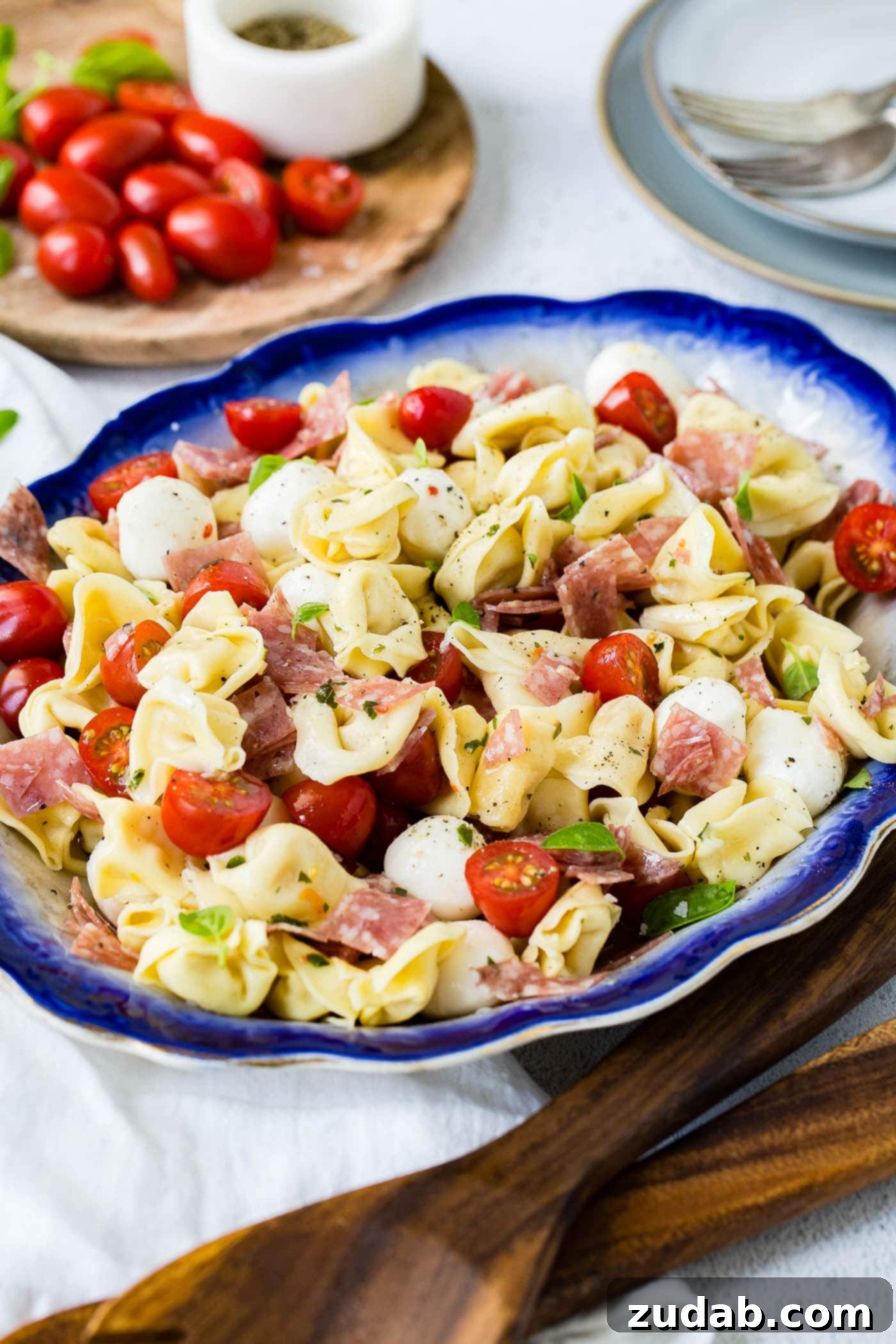 A blue-edged serving dish of 5-minute salami tortellini salad. There are chunks of savory salami, sliced grape tomatoes, and creamy mozzarella balls mixed in with the tender tortellini. A wooden cutting board with fresh grape tomatoes is in the background.