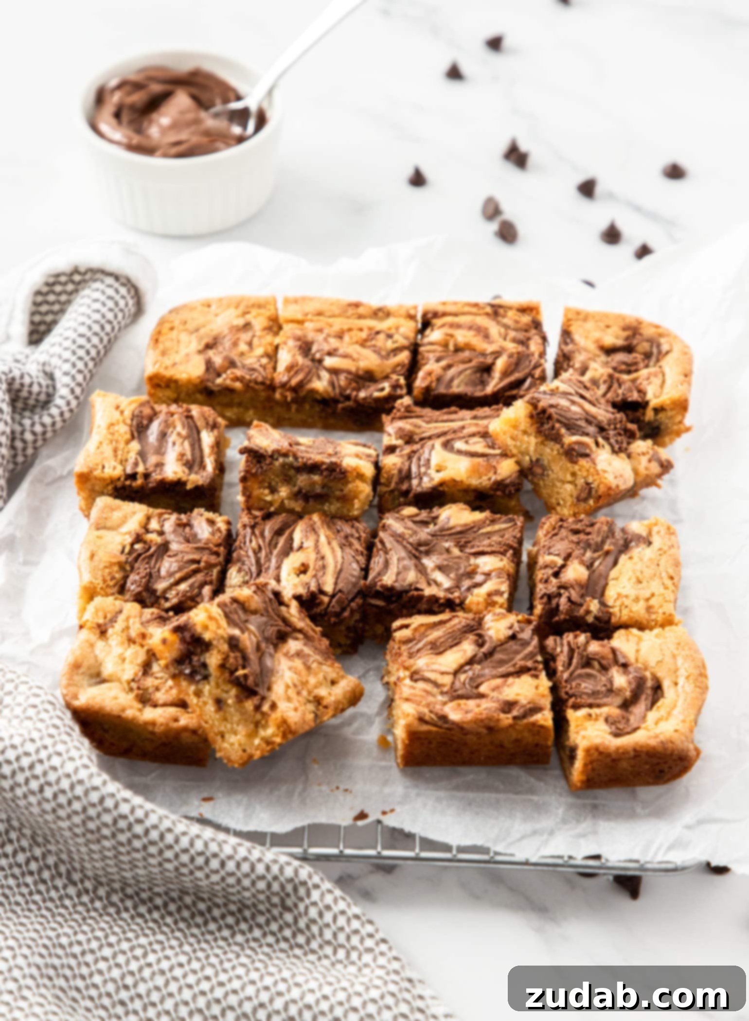 Decadent Nutella Blondies 5 A batch of blondies on a sheet on baking paper with a small bowl of nutella in the background, inviting you to bake.