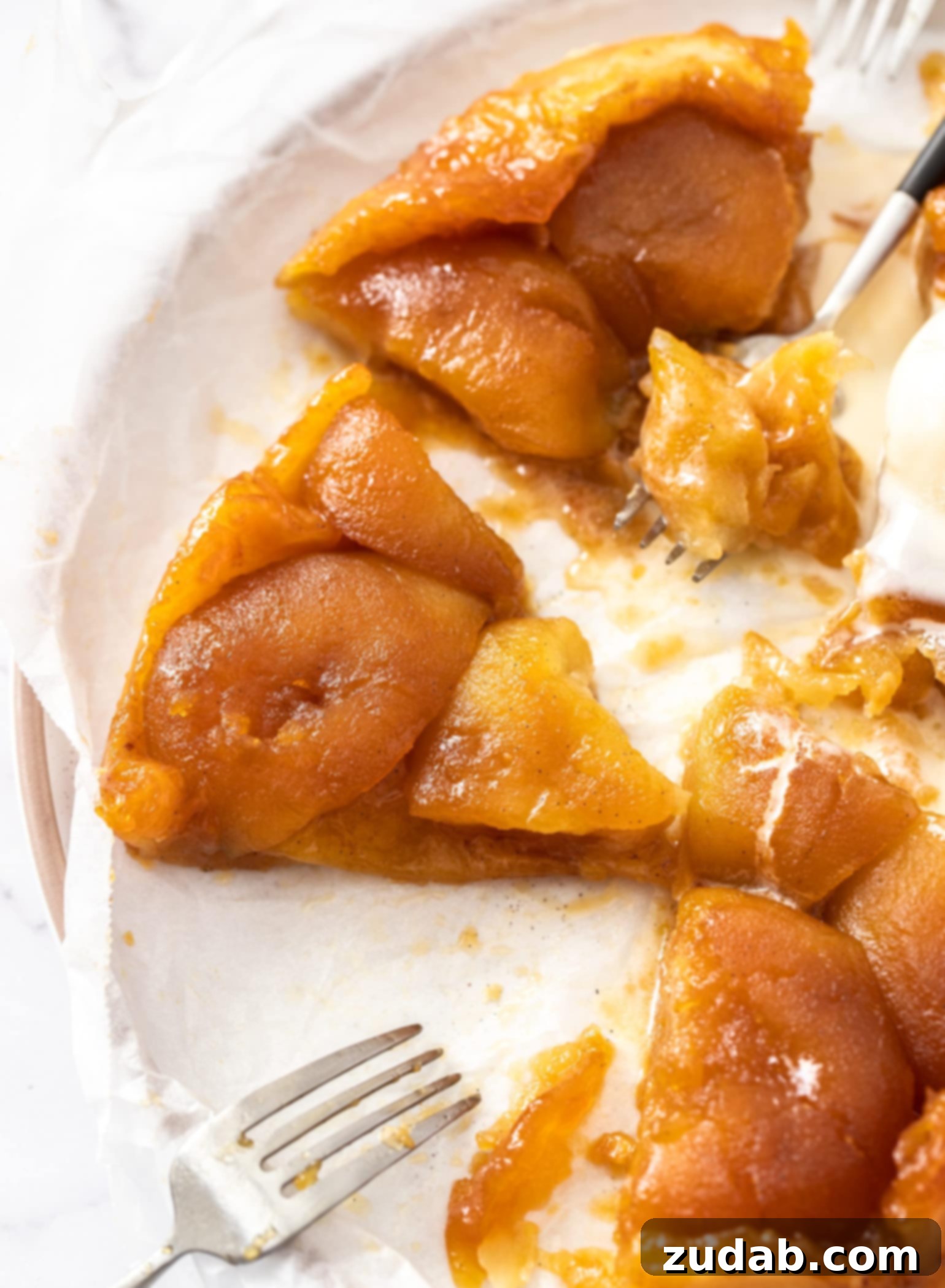 A close-up view of a perfectly baked slice of Apple Tarte Tatin on a white plate, accompanied by two dessert forks. The tender, caramelized apple slices are clearly visible beneath the crisp, golden puff pastry, promising a delightful texture and flavor. This stunning French dessert, with its sweet sticky caramel and tender apples, baked on a crispy puff pastry base, is far easier than it looks.