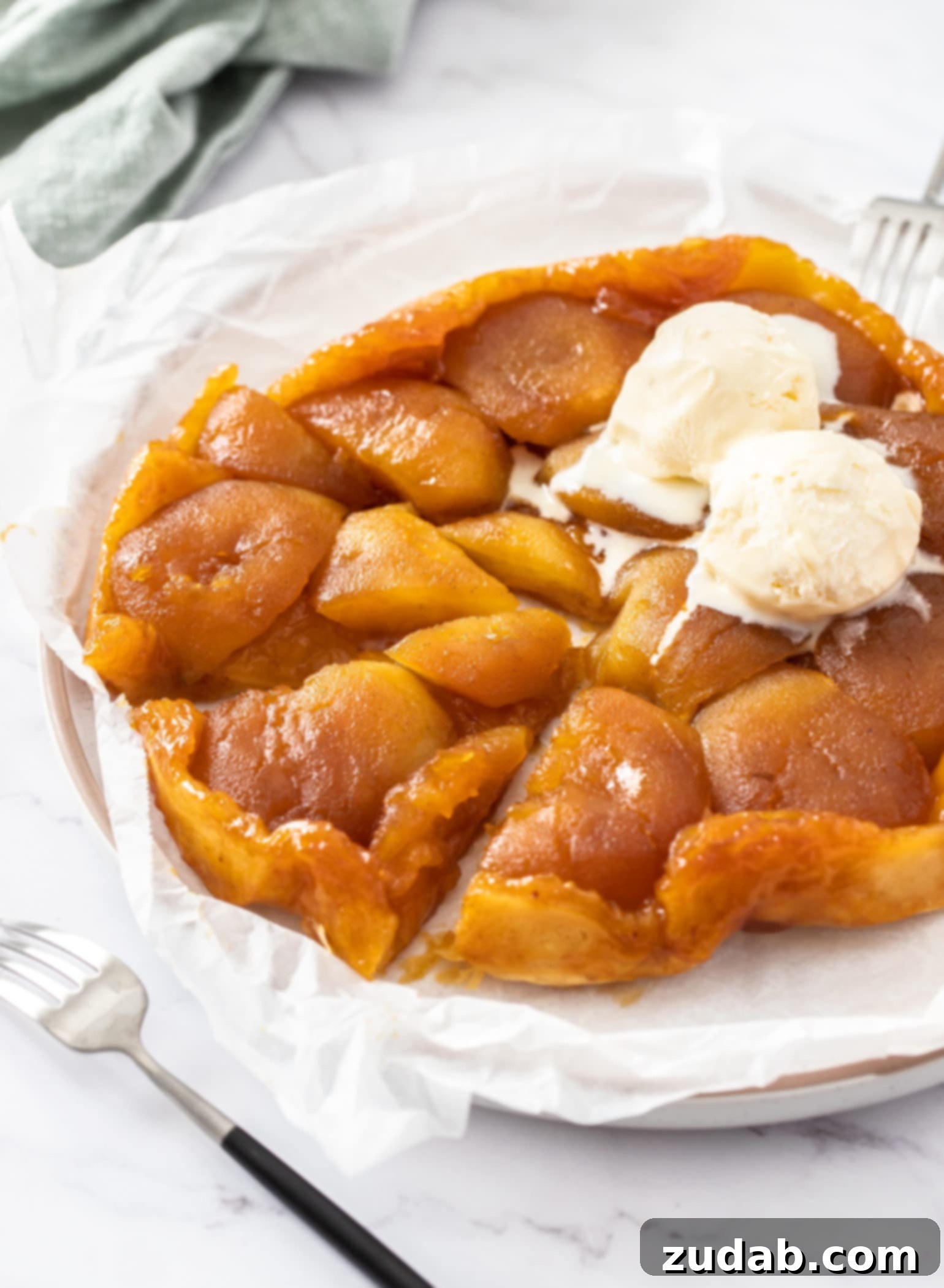 A beautifully inverted Apple Tarte Tatin, golden and glistening with caramel, sits elegantly on a sheet of baking paper on a marble surface. It's crowned with a generous scoop of creamy ice cream, ready to be served. This image captures the deliciousness and beauty of the classic French apple tart, showcasing its tender, sticky caramel apples and crispy puff pastry base.