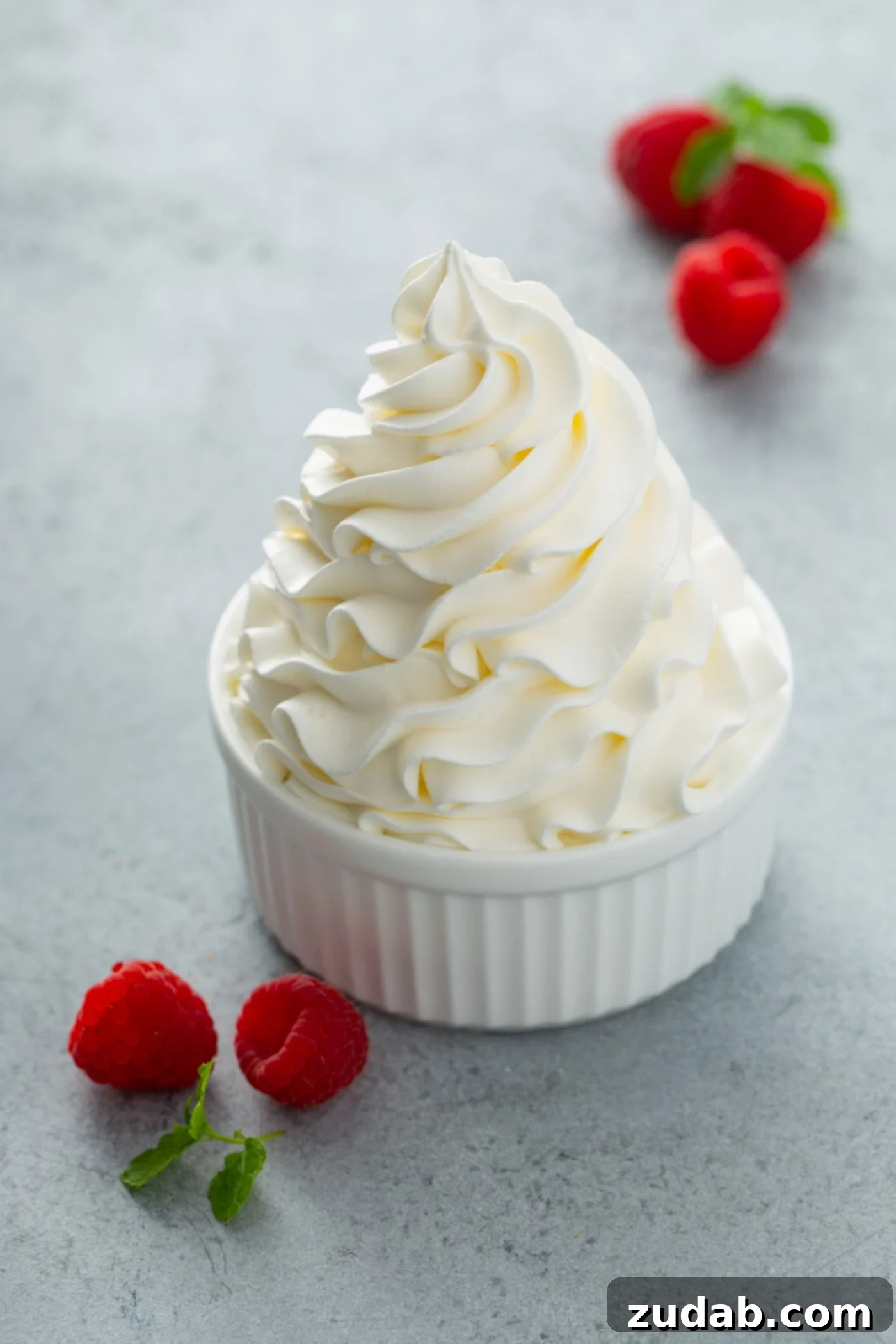 Long Lasting Whipped Cream A bowl of stabilized whipped cream that is swirled to a peak. A few raspberries are scattered around the dish.