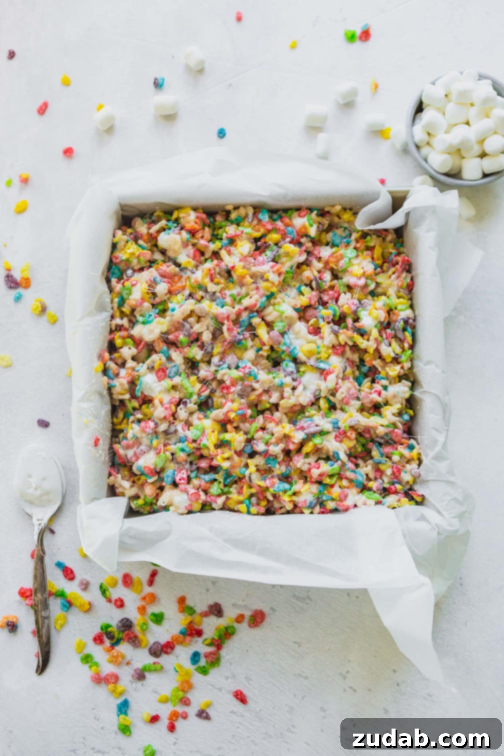 Colorful Fruity Pebbles Rice Krispie Treats A parchment-lined baking pan filled with freshly made Fruity Pebbles Rice Krispie treats, with a few loose pebbles scattered on the table.