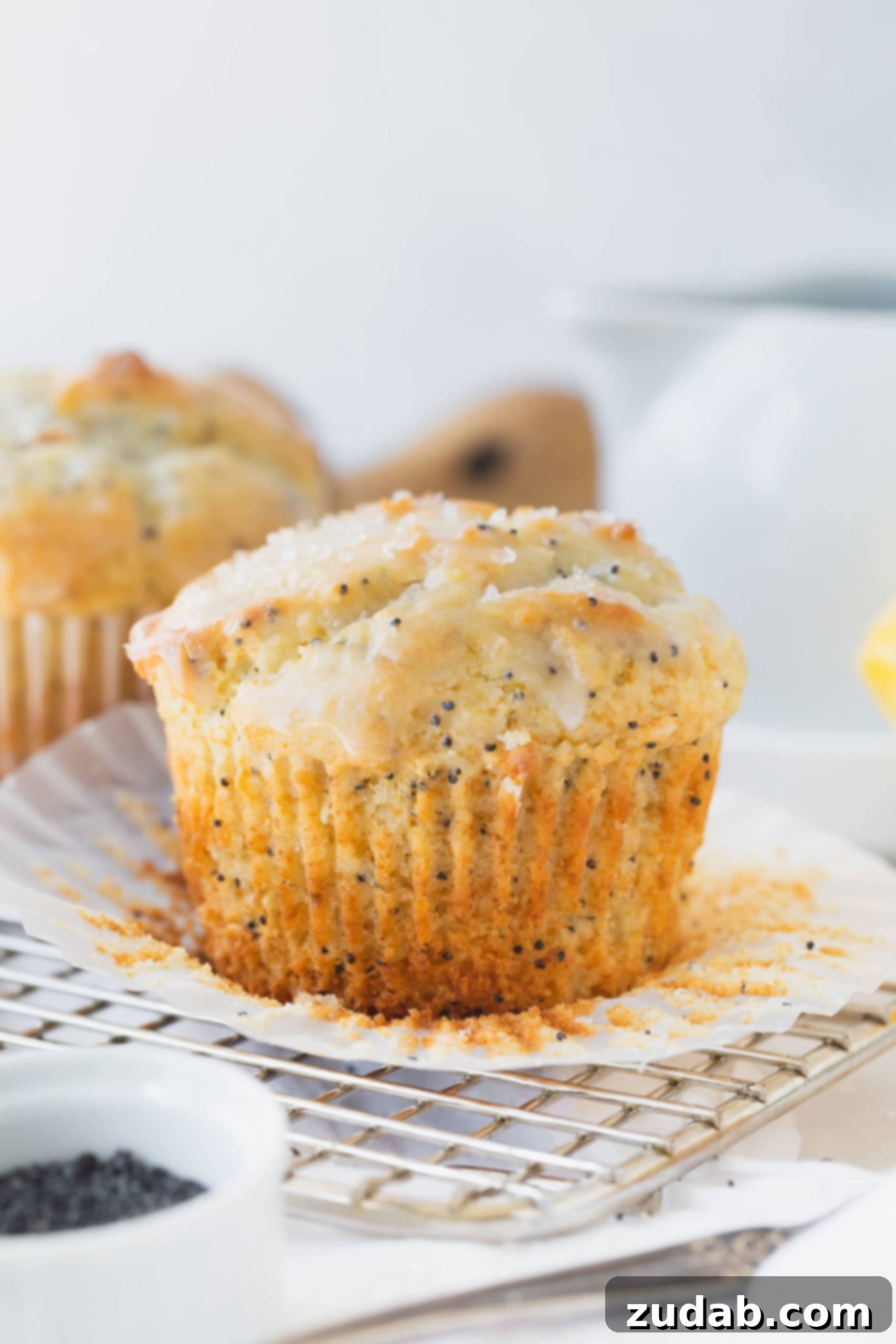 Lemon Poppy Seed Muffins A lemon glazed lemon poppy seed muffin with the paper liner peeled back. there is another muffin in the background.