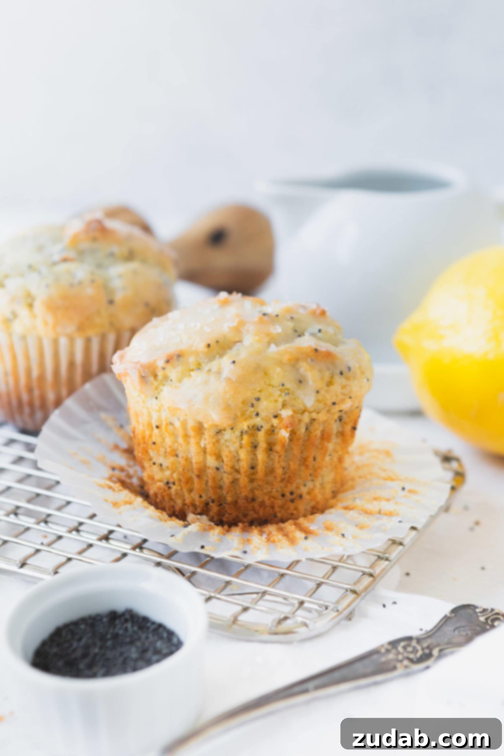 Lemon Poppy Seed Muffins A lemon poppy seed muffin that has the paper liner peeled back. The muffin is glazed with a lemon glaze and there is another muffin and a lemon, a container of poppy seeds in the background.