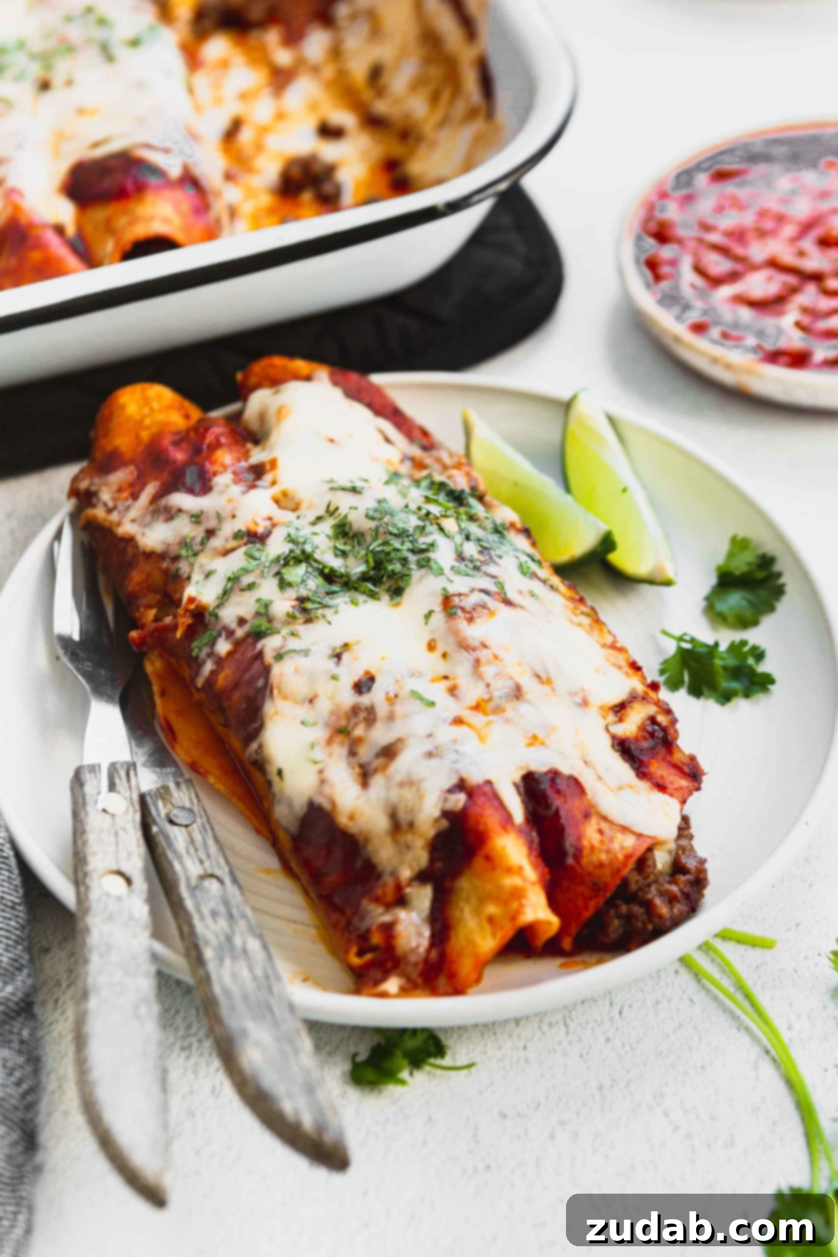 Family Favorite Cheesy Ground Beef Enchiladas A photo of two ground beef enchiladas on a white plate with two lime wedges on the side.