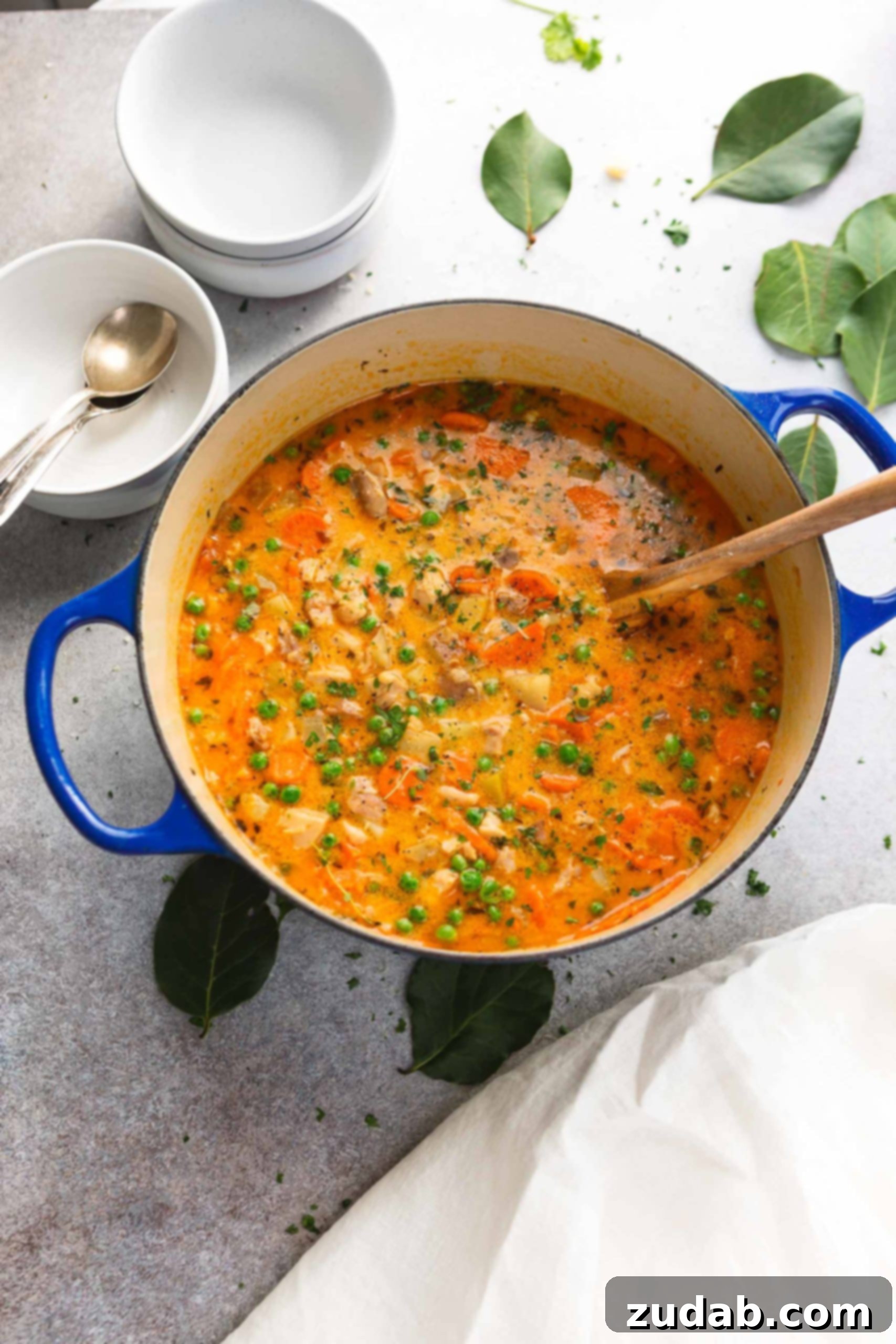 Hearty Slow Cooker Chicken Stew 2 A pot of slow cooker chicken stew with a wooden spoon. The stew chicken and vegetables are in a thick yellow-red gravy and there are bright green herbs with the vegetables. A stack of bowls and spoons are in the background.