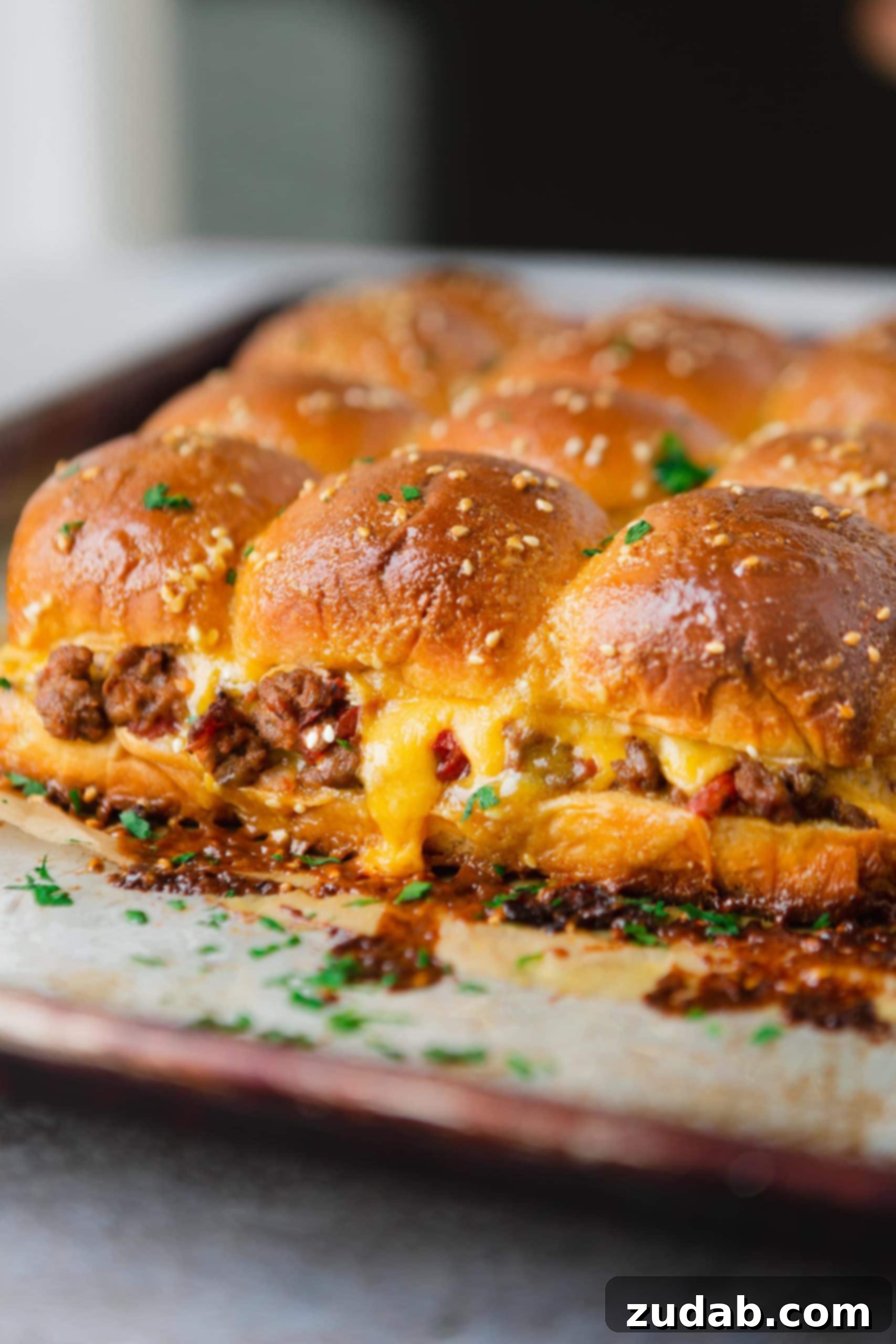 A panful of cheeseburger sliders. The cheese is oozing from the rolls and you can see ground beef and chunks of tomatoes.