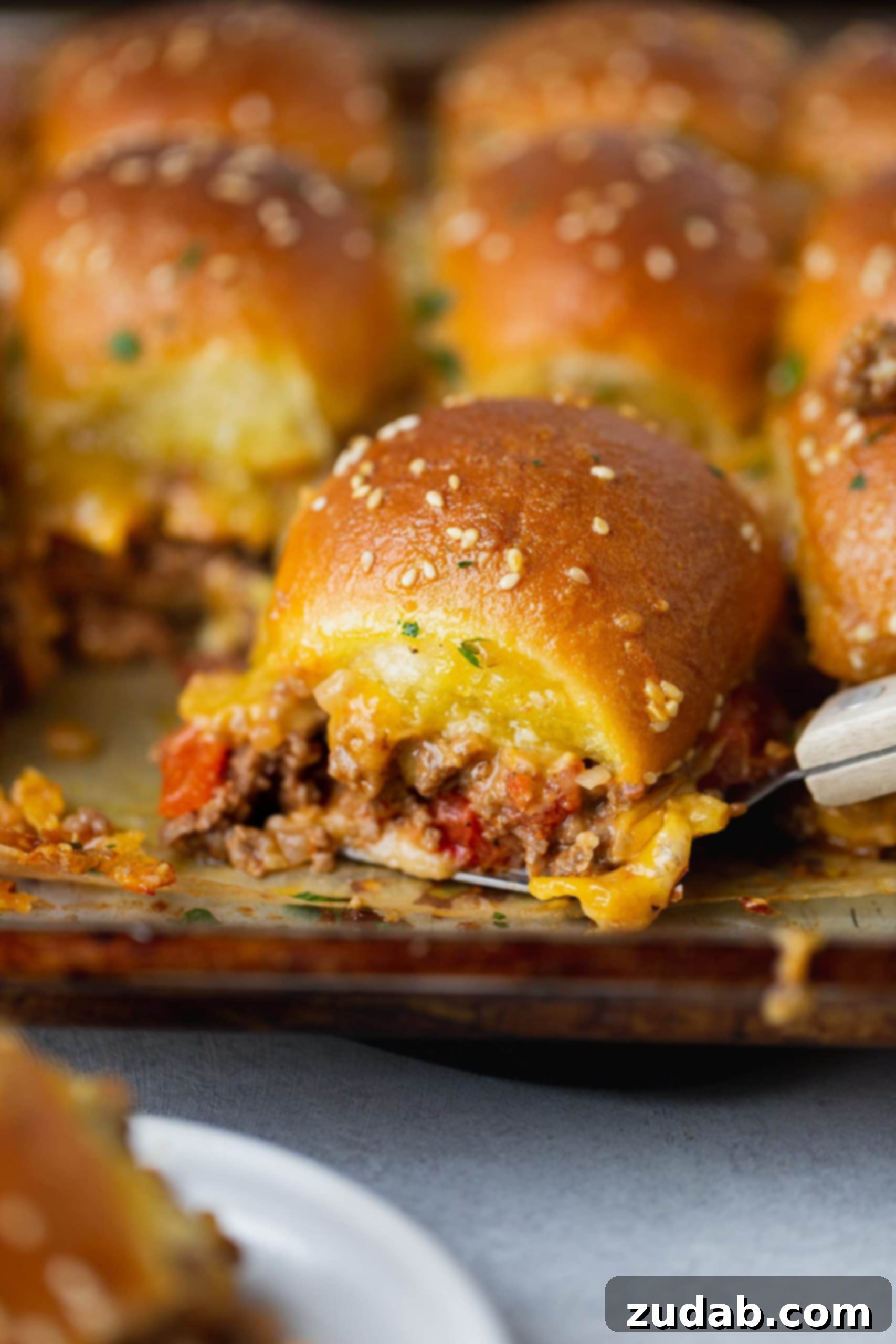 A cheeseburger slider filled with ground beef, melty cheese and chopped tomatoes. there are other sliders in the background.