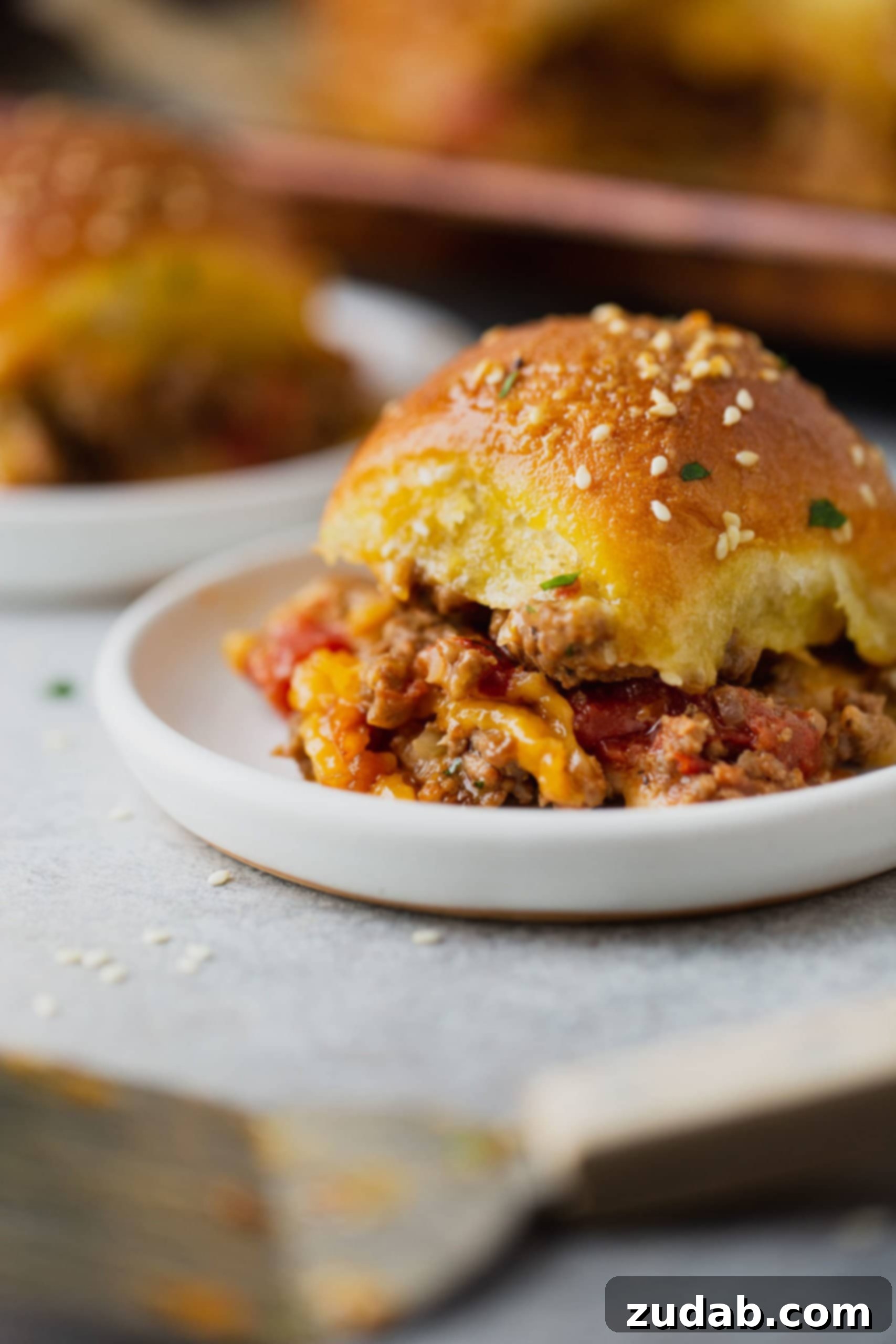 A cheeseburger slider on a plate. The golden roll is packed with ground beef, melted cheese and copped tomatoes.
