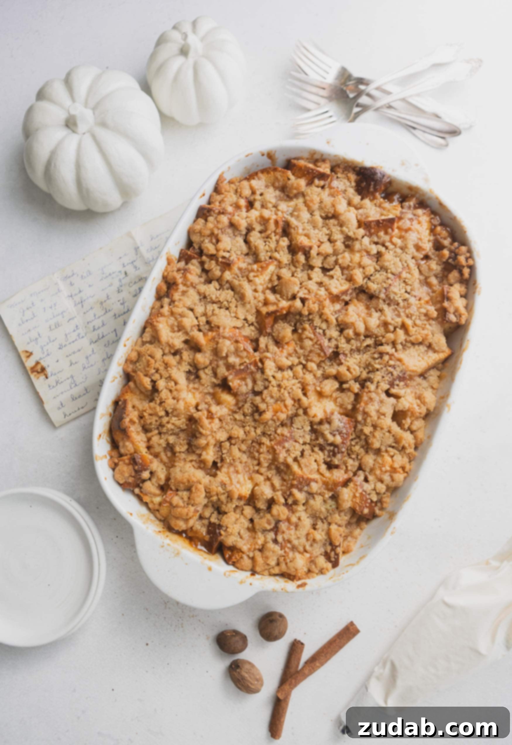 Eggnog French Toast Casserole A casserole dish of eggnog french toast. It is topped with a brown crumb topping. A small white pumpkin is in the background.