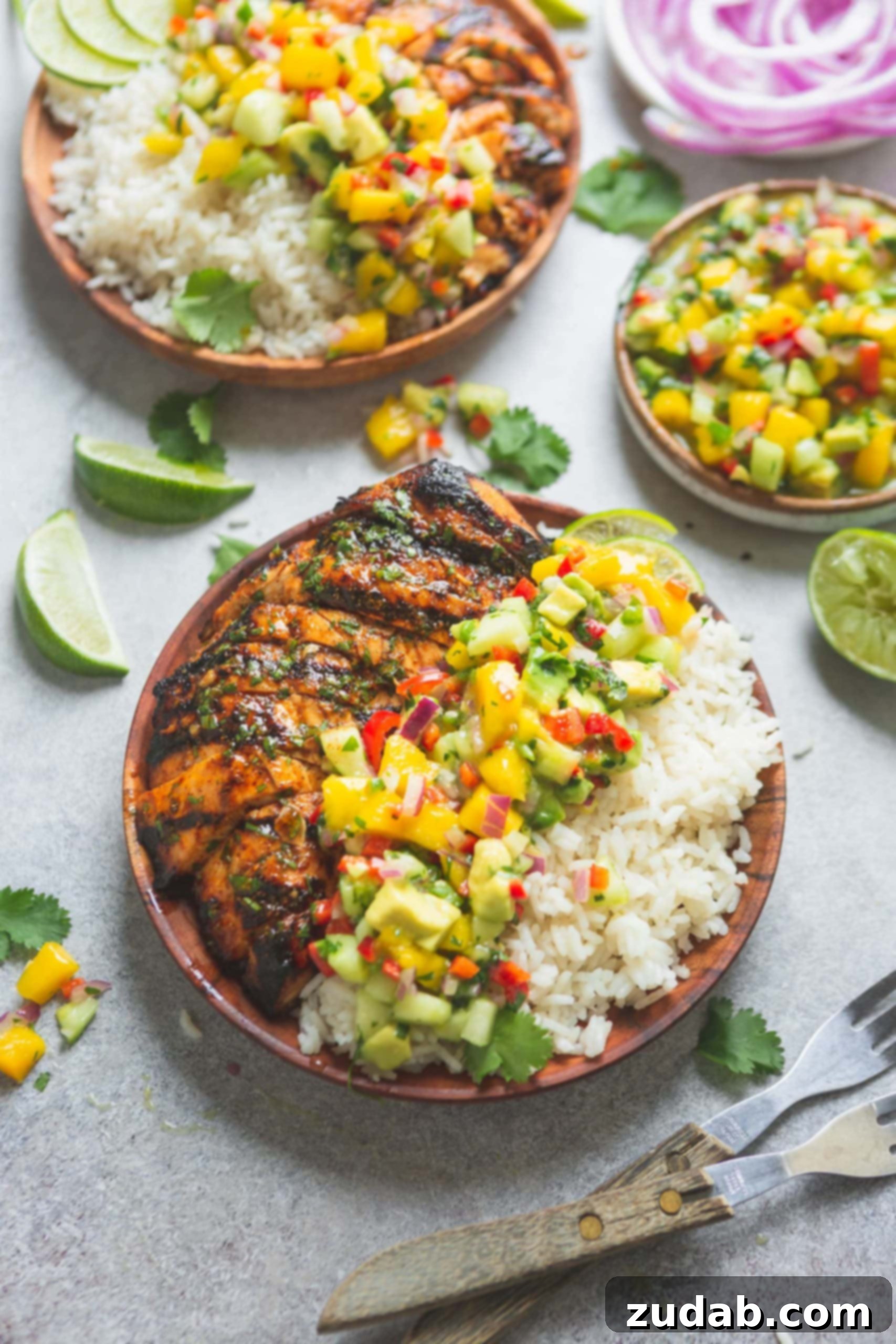 A plate filled with perfectly grilled Mexican chicken, fluffy coconut rice, and a generous topping of bright, colorful mango salsa. The salsa features visible chunks of mango, vibrant peppers, and fresh cilantro, garnished with lime wedges. Other plates and ingredients are softly blurred in the background, suggesting a festive meal.