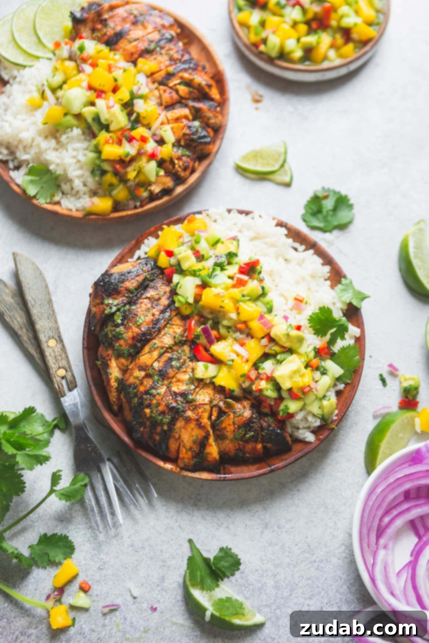 Two vibrant dinner plates showcase grilled chicken, coconut rice, and a generous portion of mango salsa, bursting with colorful chunks of mango, red onion, bell peppers, and fresh cilantro. Sliced red onions, lime wedges, and forks are arranged on the table, inviting diners to enjoy this fresh and appealing meal.