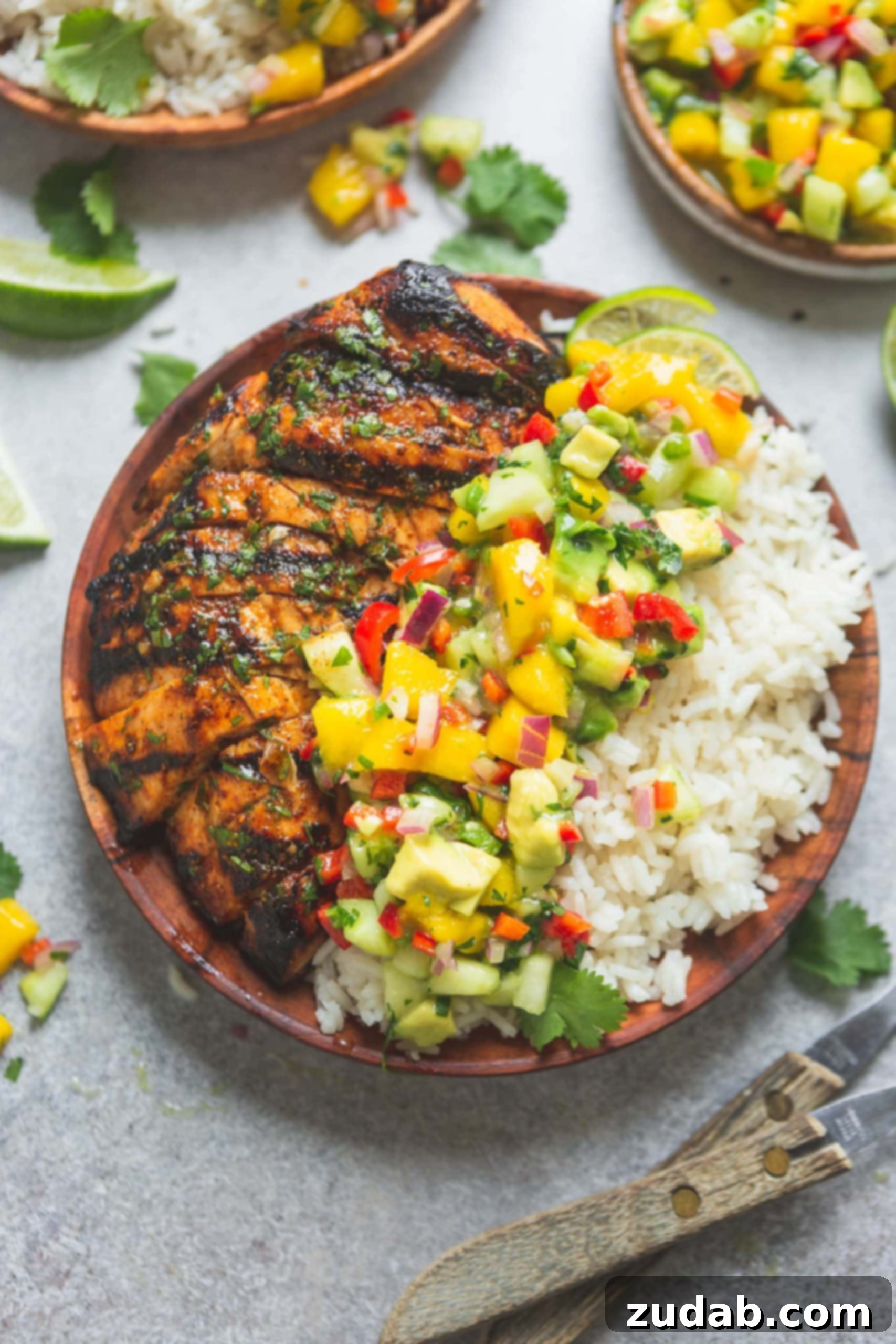 A dinner plate piled high with golden-brown grilled chicken and fragrant coconut rice, generously topped with a vibrant mango salsa. The salsa is a colorful mix of diced mango, various peppers, red onions, and fresh cilantro, showcasing the freshness of the ingredients.