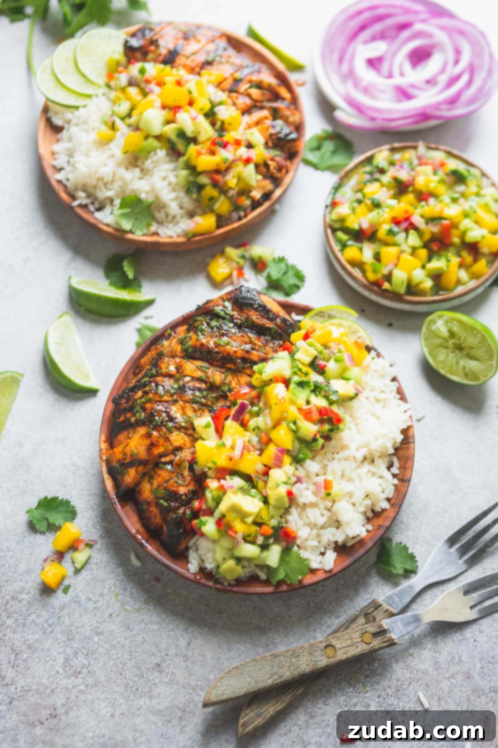 A beautifully presented dinner plate laden with grilled Mexican chicken, a mound of creamy coconut rice, and a colorful topping of mango salsa. The salsa is bright with distinct pieces of mango, peppers, and cilantro. In the background, a bowl of fresh salsa and another plate with the full meal are visible, alongside scattered lime wedges on the table, enhancing the fresh and inviting scene.