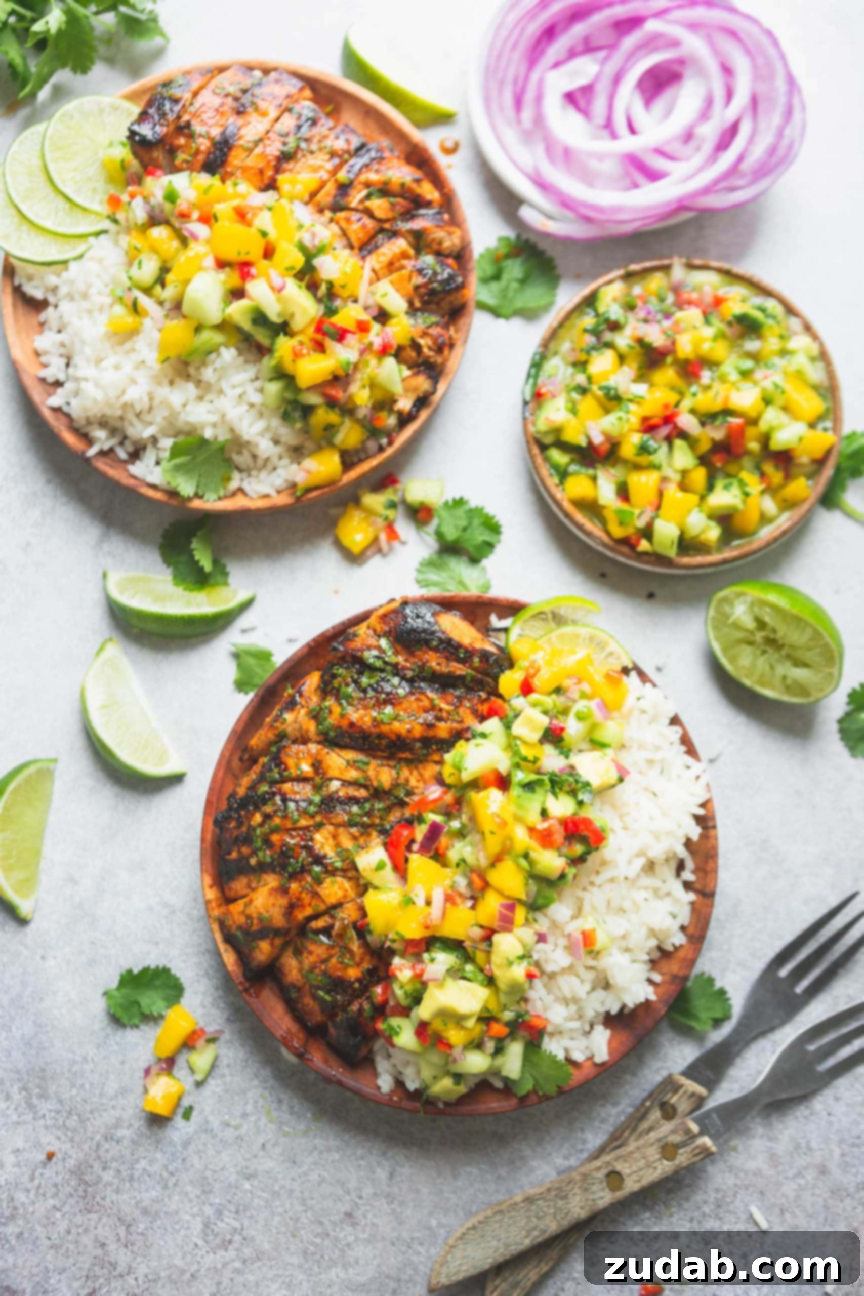 Two plates generously filled with grilled Mexican chicken, creamy coconut rice, and a vibrant mango salsa, which features large chunks of mango, colorful peppers, and fresh cilantro. A separate bowl of the fresh salsa is on the table, surrounded by scattered lime wedges and fresh cilantro leaves, with two forks indicating readiness to enjoy.