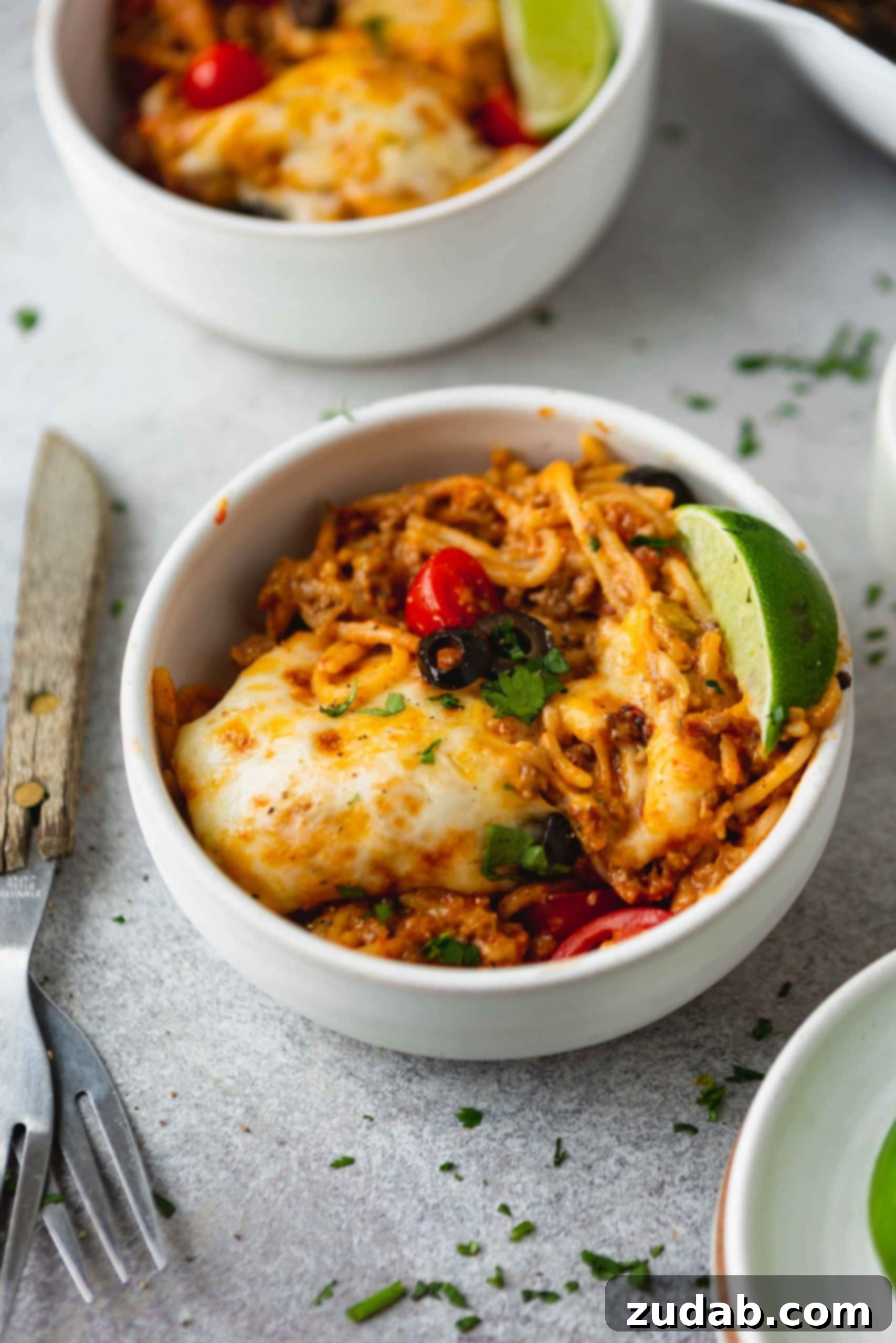 One Pot Cheesy Ground Beef Taco Spaghetti A bowl of cheesy ground beef taco spaghetti. It has spaghetti, ground beef, cheese, green onions, tomatoes, olives and cilantro. A lime wedge is on the edge of the bowl and two forks are near the bowl.