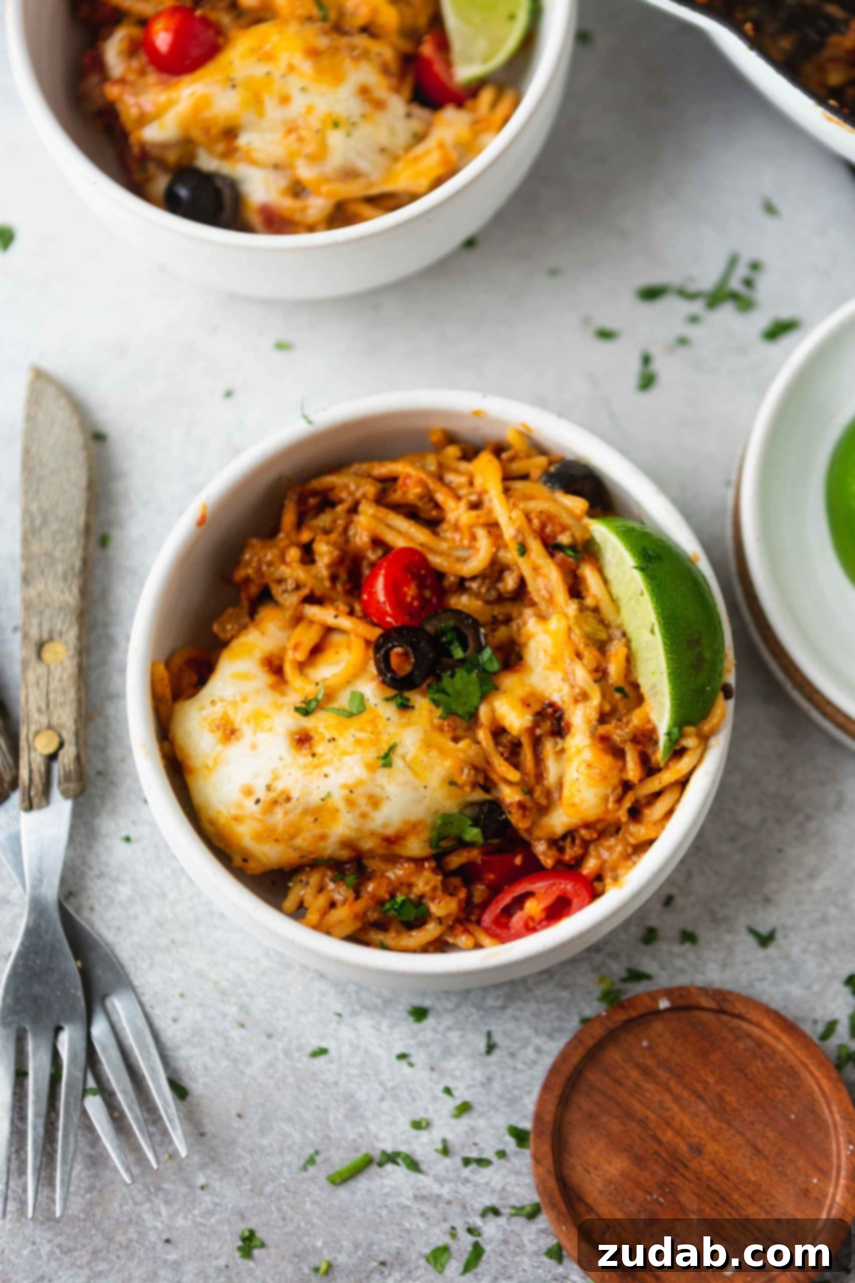 One Pot Cheesy Ground Beef Taco Spaghetti A dinner bowl of cheesy ground beef taco spaghetti. There is a lime wedge on the edge of the bowl. The spaghetti is topped with melted cheese, ground beef, green onions, olives and tomatoes. Two forks are next to the bowl.