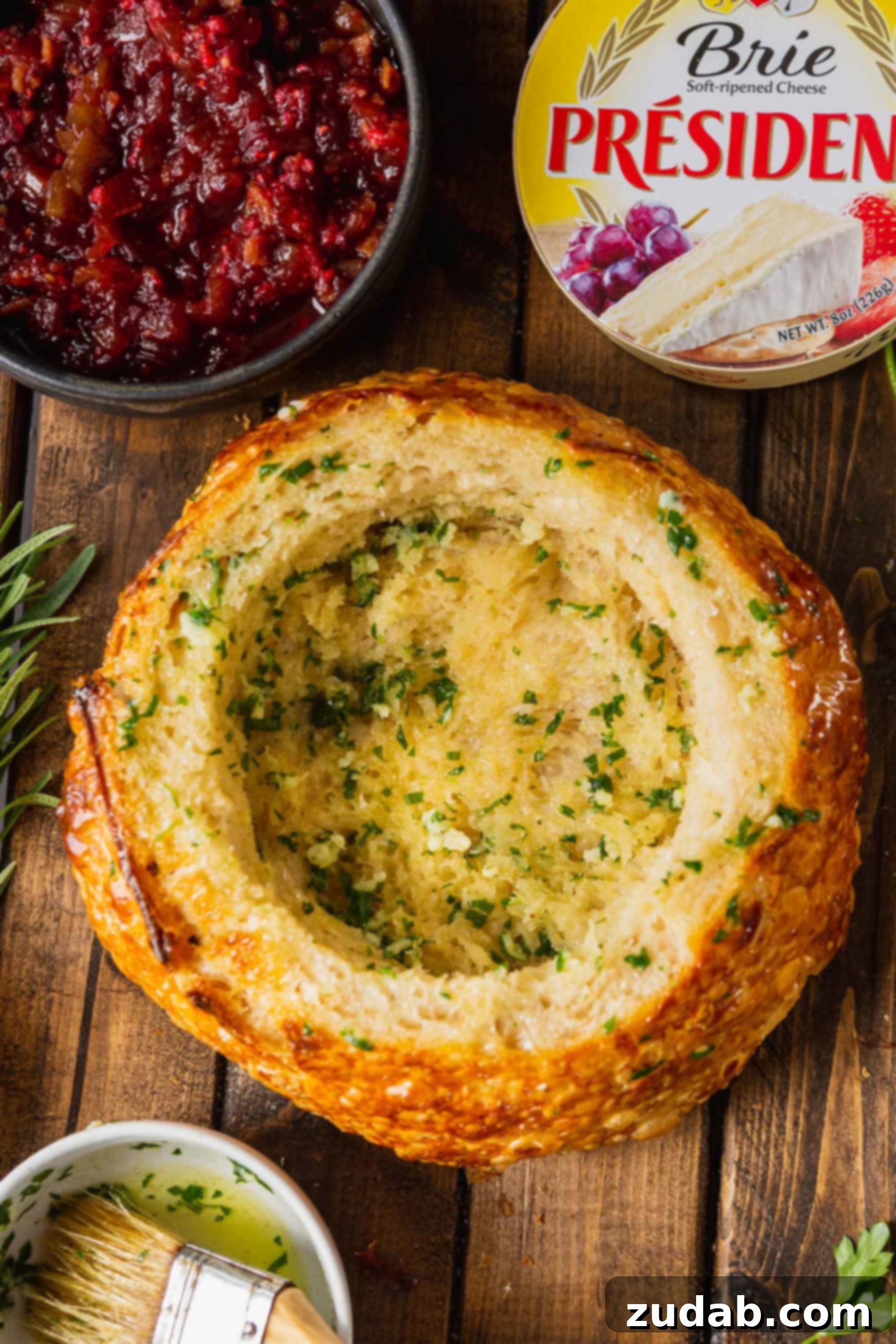 The Most Incredible Pull Apart Baked Brie A freshly baked sourdough bread bowl, brimming with warm, melted brie cheese and topped with a vibrant raspberry bacon onion jam, garnished with fresh herbs.