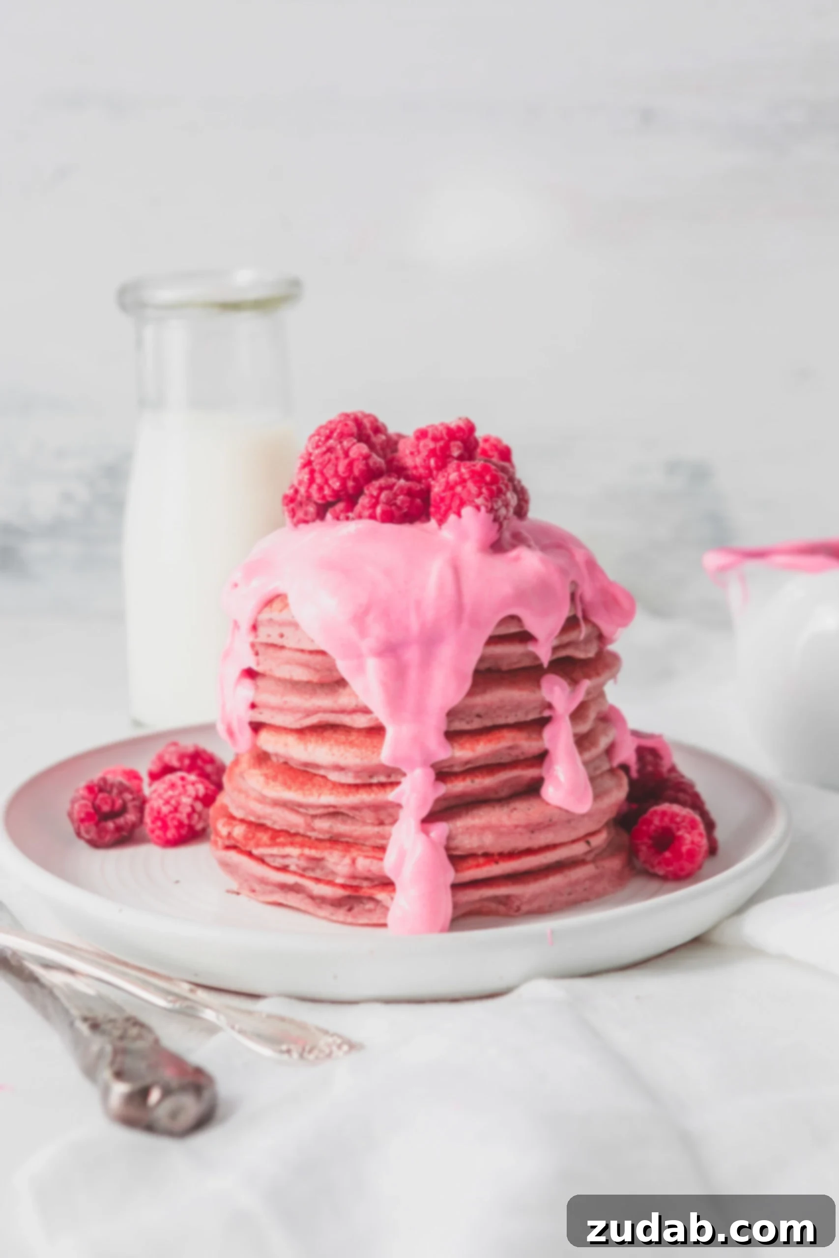 Healthy Pink Pitaya Blender Pancakes A large stack of healthy pink pitaya blender pancakes, generously covered with a thick pink chocolate sauce. Fresh raspberries are elegantly placed on top of the sauce and scattered around the plate, enhancing the visual appeal and natural sweetness.