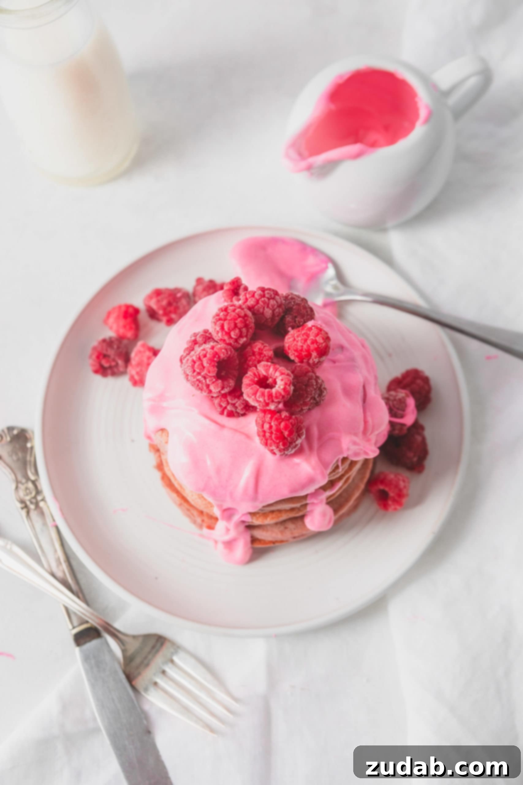 Healthy Pink Pitaya Blender Pancakes An overhead view of a stack of healthy pink pitaya blender pancakes on a pristine white plate. The pancakes are generously adorned with a thick, luscious pink chocolate sauce, and crowned with fresh, vibrant raspberries. A spoon holds a dollop of the pink sauce on the plate, while a container brimming with sauce sits invitingly in the background, ready for more.