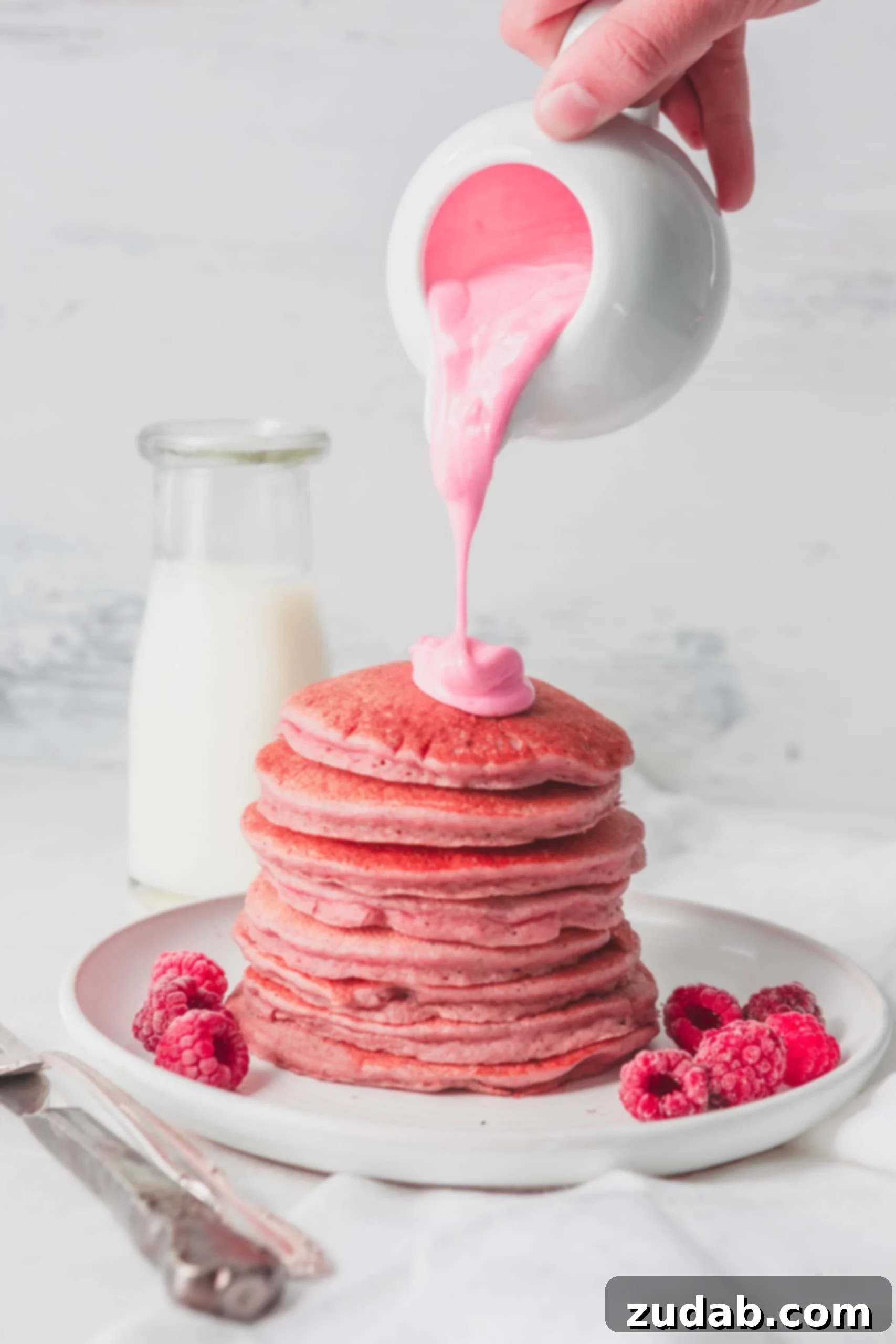 Healthy Pink Pitaya Blender Pancakes A stack of healthy pink pitaya blender pancakes, covered with a thick pink chocolate sauce being poured over the top. Fresh raspberries are elegantly arranged on the plate, while a container of milk sits in the background, adding to the delightful breakfast scene.