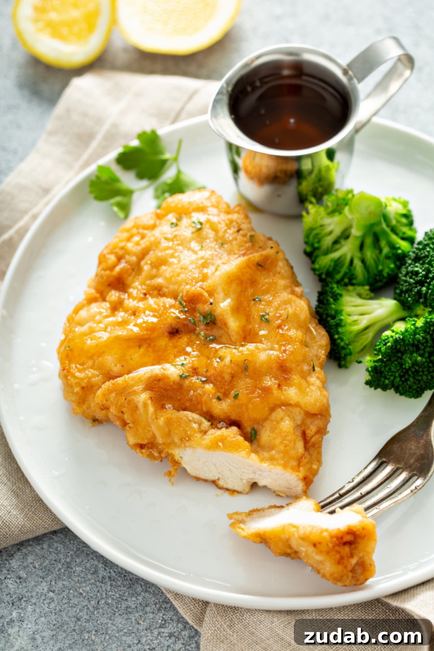 A white dinner plate containing golden brown fried chicken breast with broccoli and a silver container of dipping sauce on the side. There is a fork with a slice of chicken on it, resting on the plate.