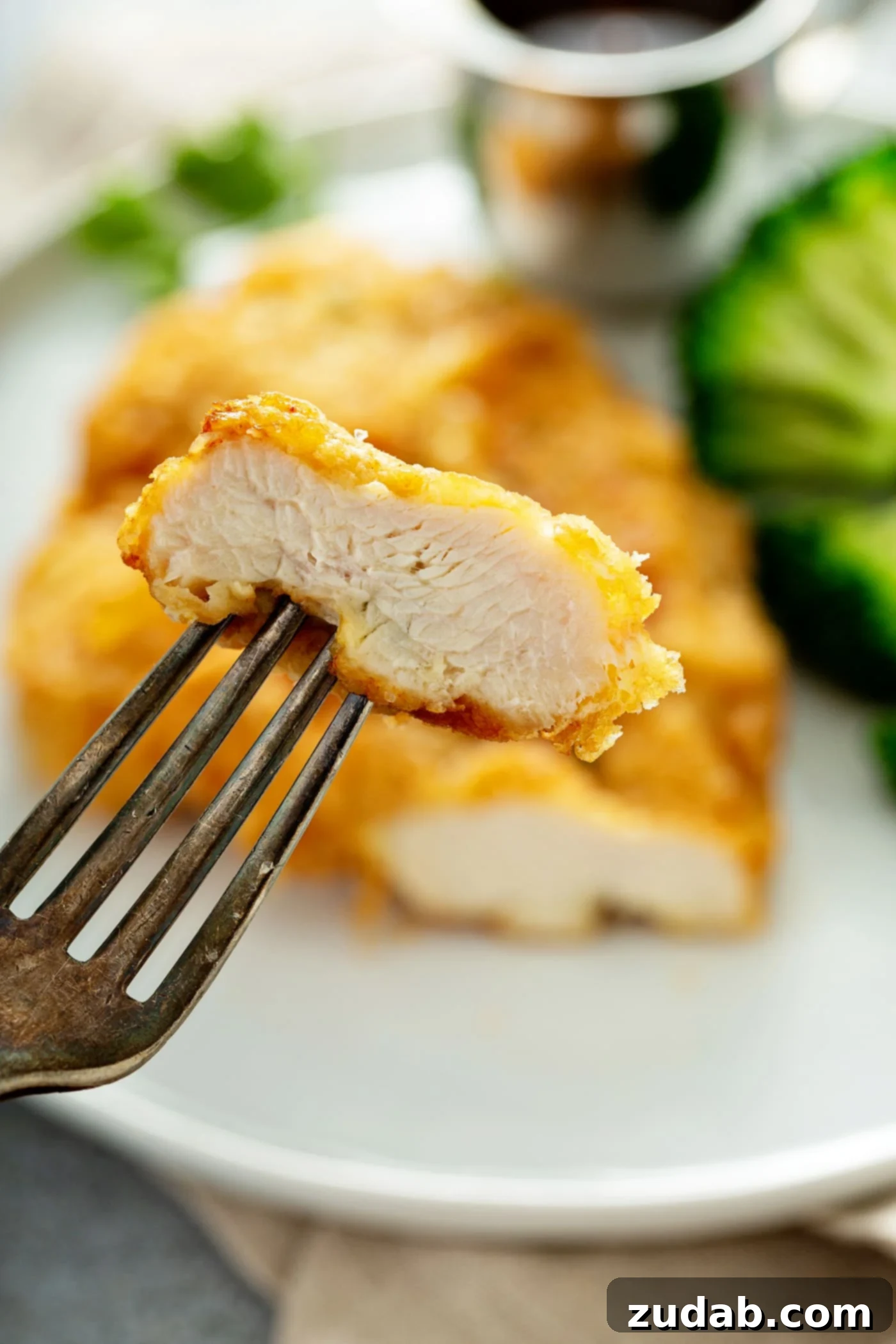 A golden brown fried chicken breast with a slice cut off and held up in the tines of a fork. There is broccoli beside the chicken on the plate.
