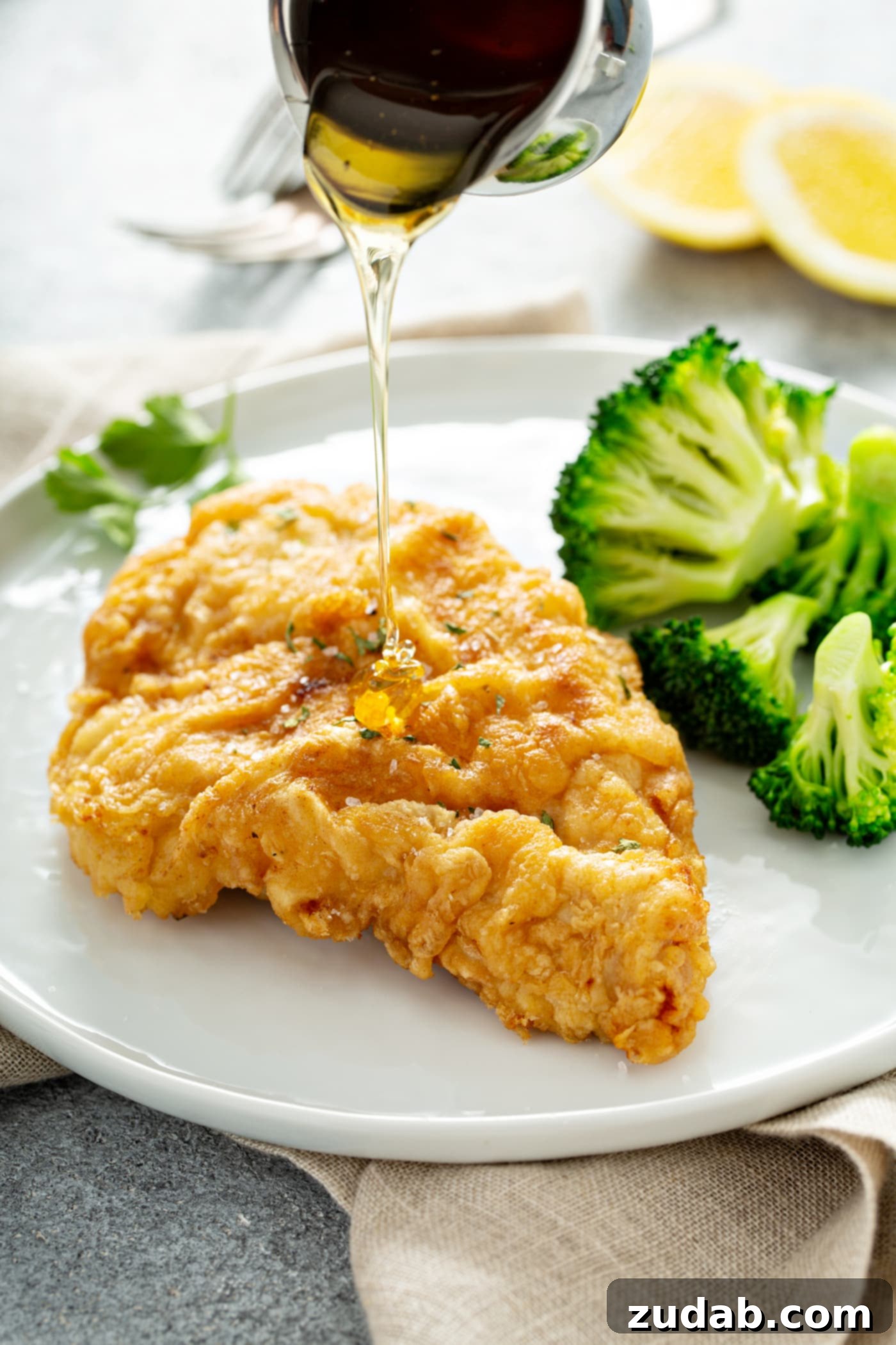 A chicken breast that has been fried to a golden brown, resting on a white dinner plate. There is steamed broccoli next to the chicken, and a sauce is being drizzled over the chicken.
