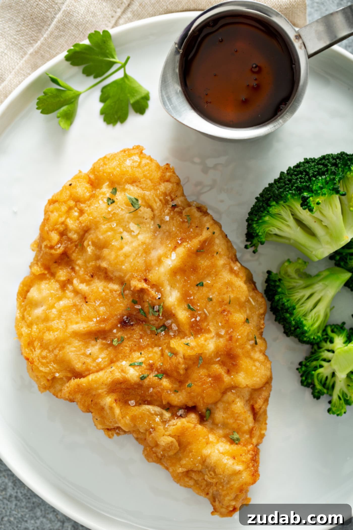 A chicken breast that has been fried to a golden brown, resting on a white dinner plate. There is also steamed broccoli next to the chicken and a small pitcher of sauce.