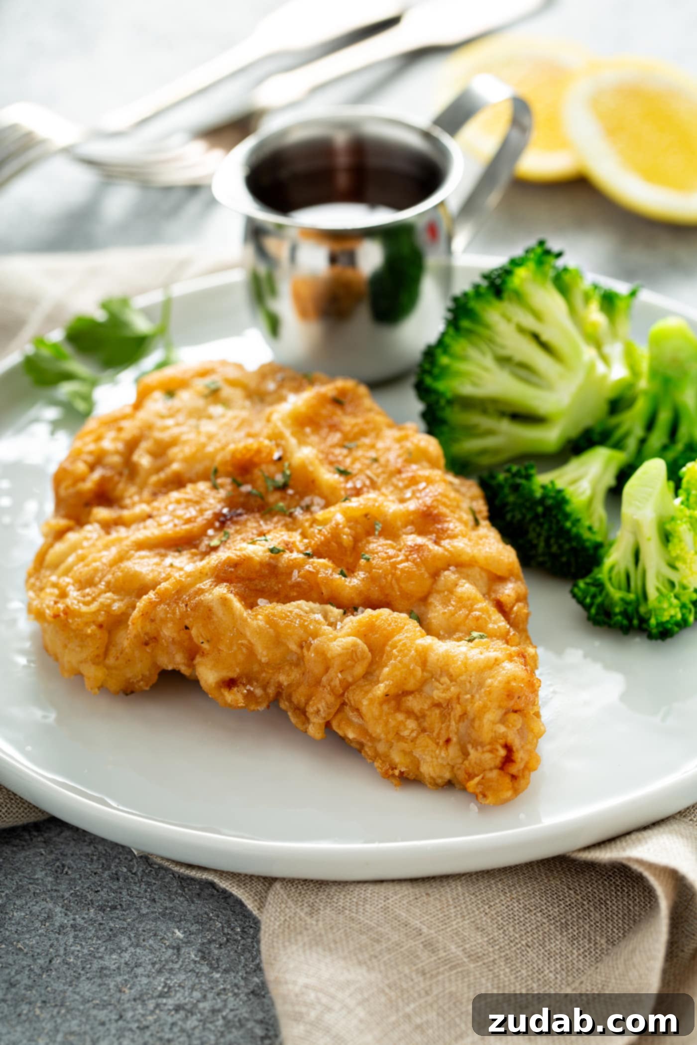 A piece of chicken fried to a perfect golden brown. There is a silver pitcher of sauce and steamed broccoli next to the chicken on a white dinner plate.
