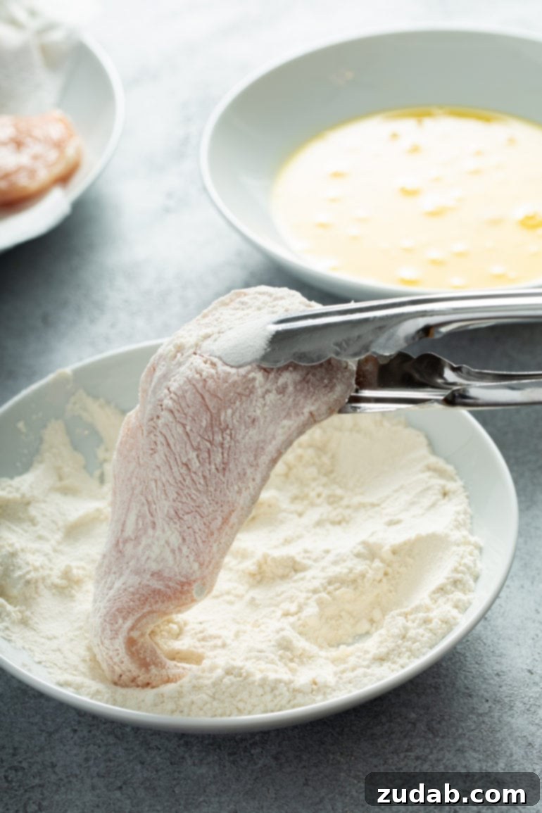 A chicken breast held with tongs, being dredged in eggs and flour.