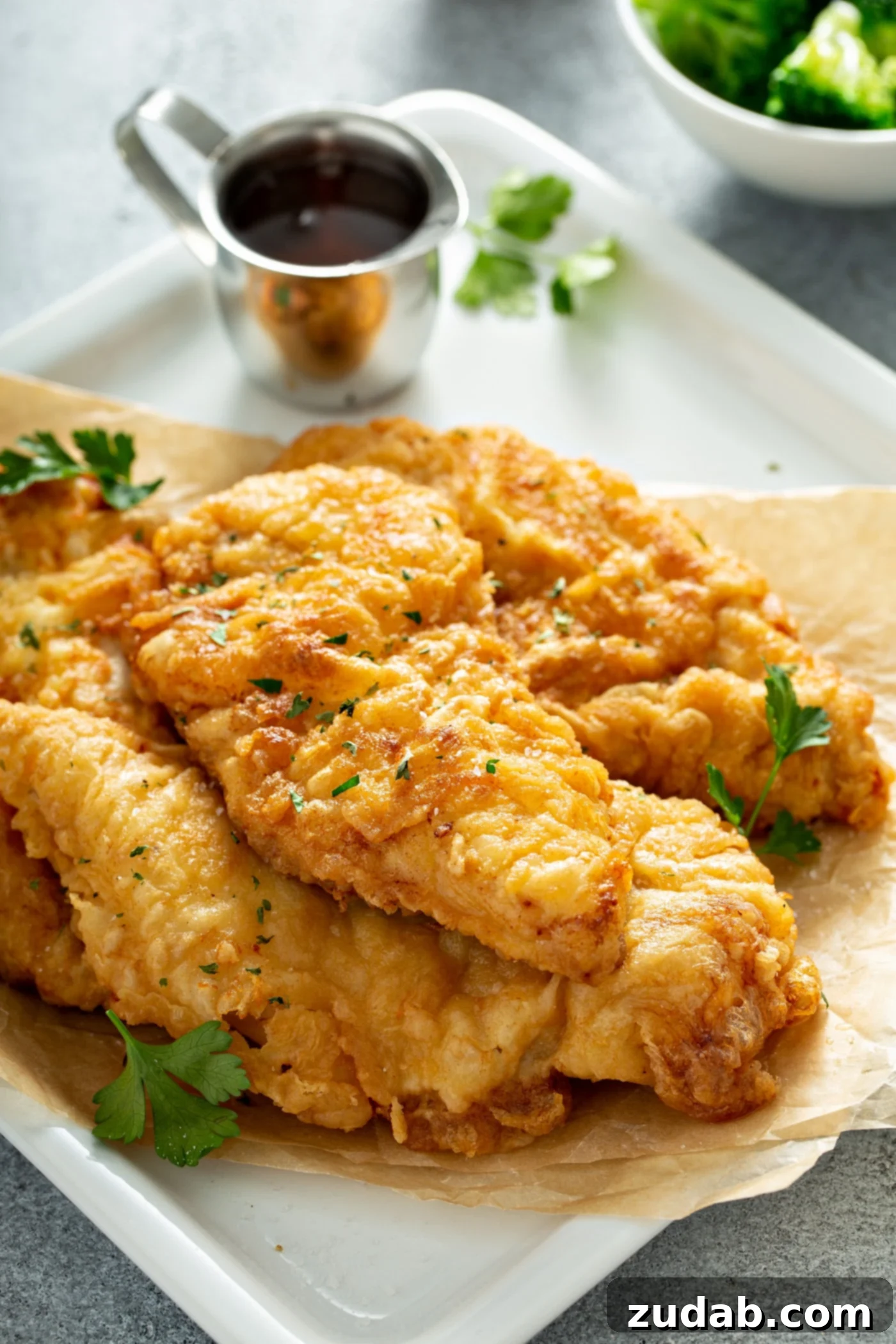 A serving platter heaped with golden fried chicken. There is a small silver pitcher of honey in the background and the platter is sprinkled with parsley leaves.