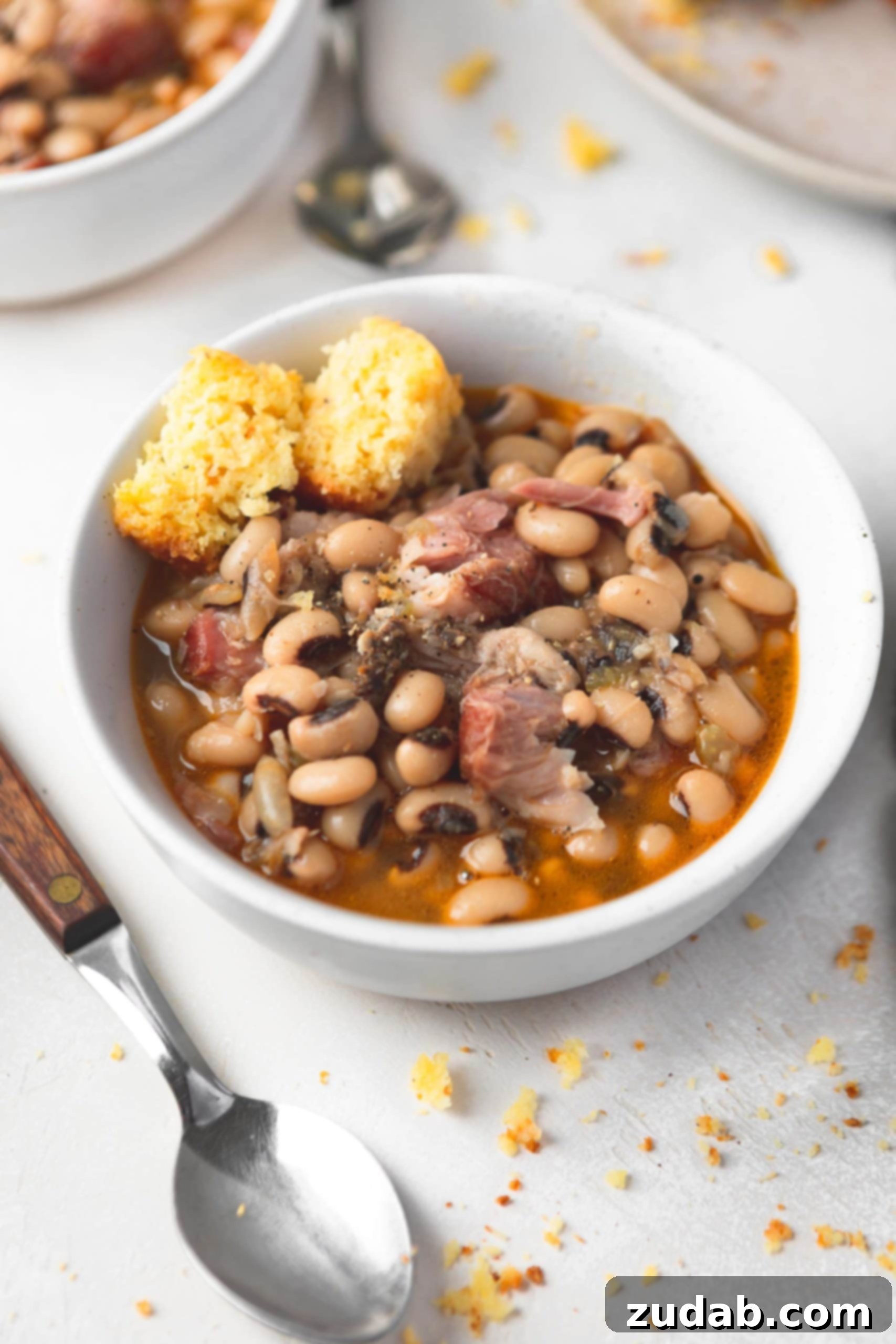 A bowl of southern black eyed peas. There are chunks of ham in the peas and a broken piece of cornbread on the side.